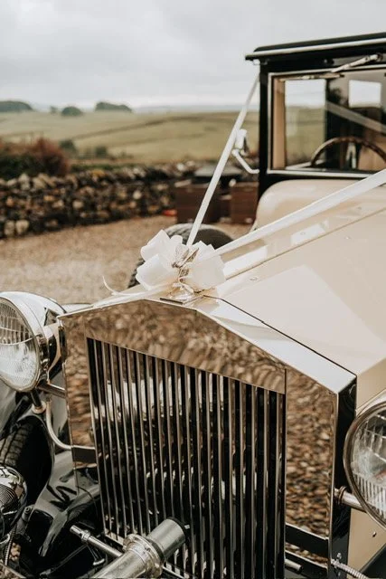 Vintage car with wedding decorations and ribbons on the hood, parked outdoors on a gravel path, with a countryside background.