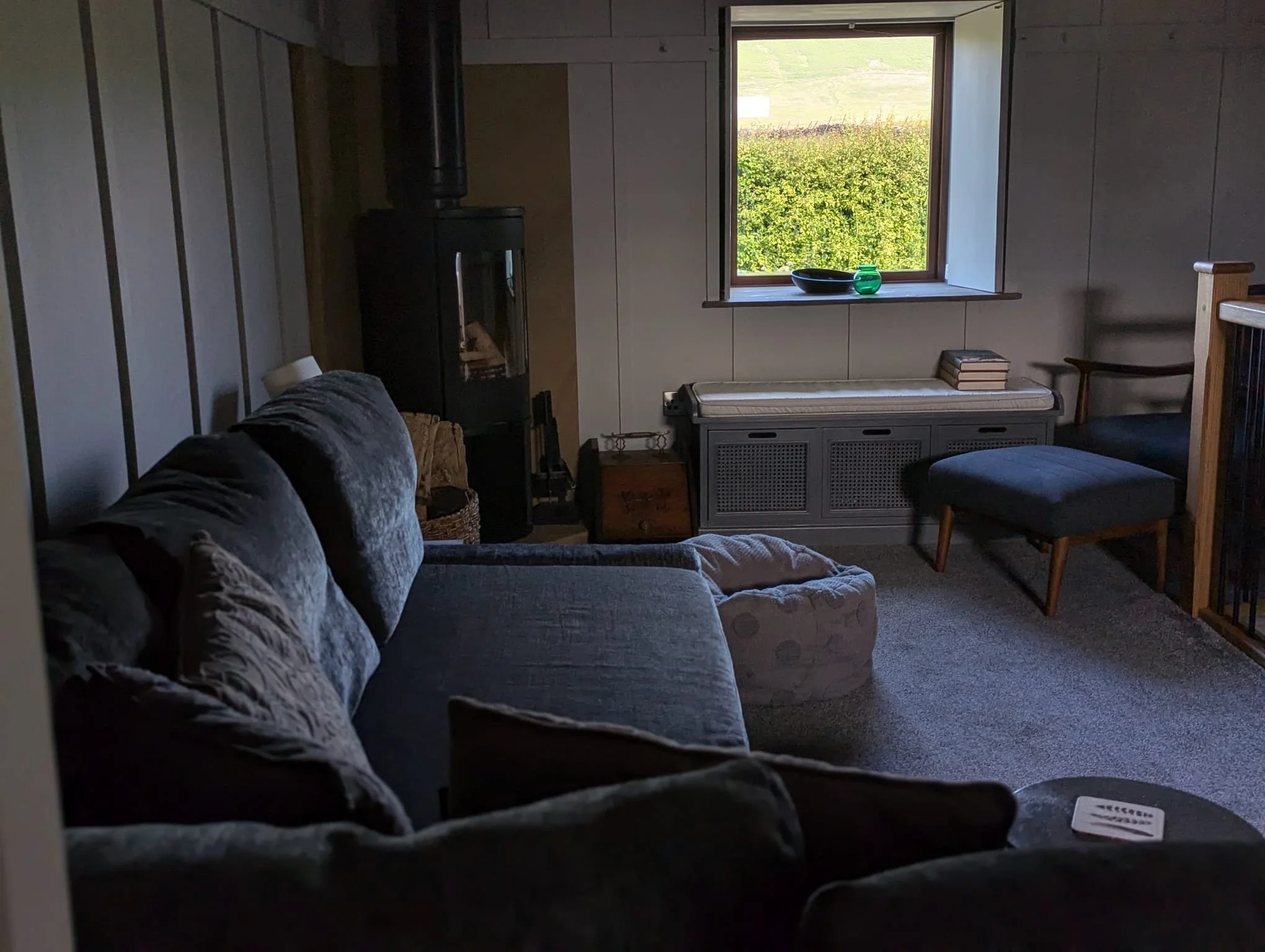 Cozy living room with dark gray sofa, wood-burning stove, and window view of greenery.