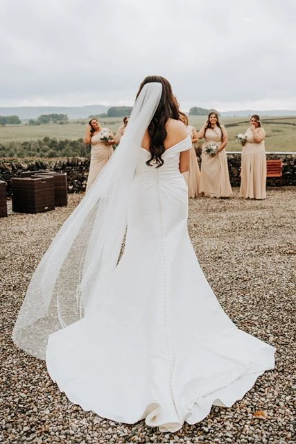 Bride in a white wedding dress with a long train and veil stands on gravel facing a group of four bridesmaids in matching dresses holding bouquets on a cloudy day with greenery in the background.