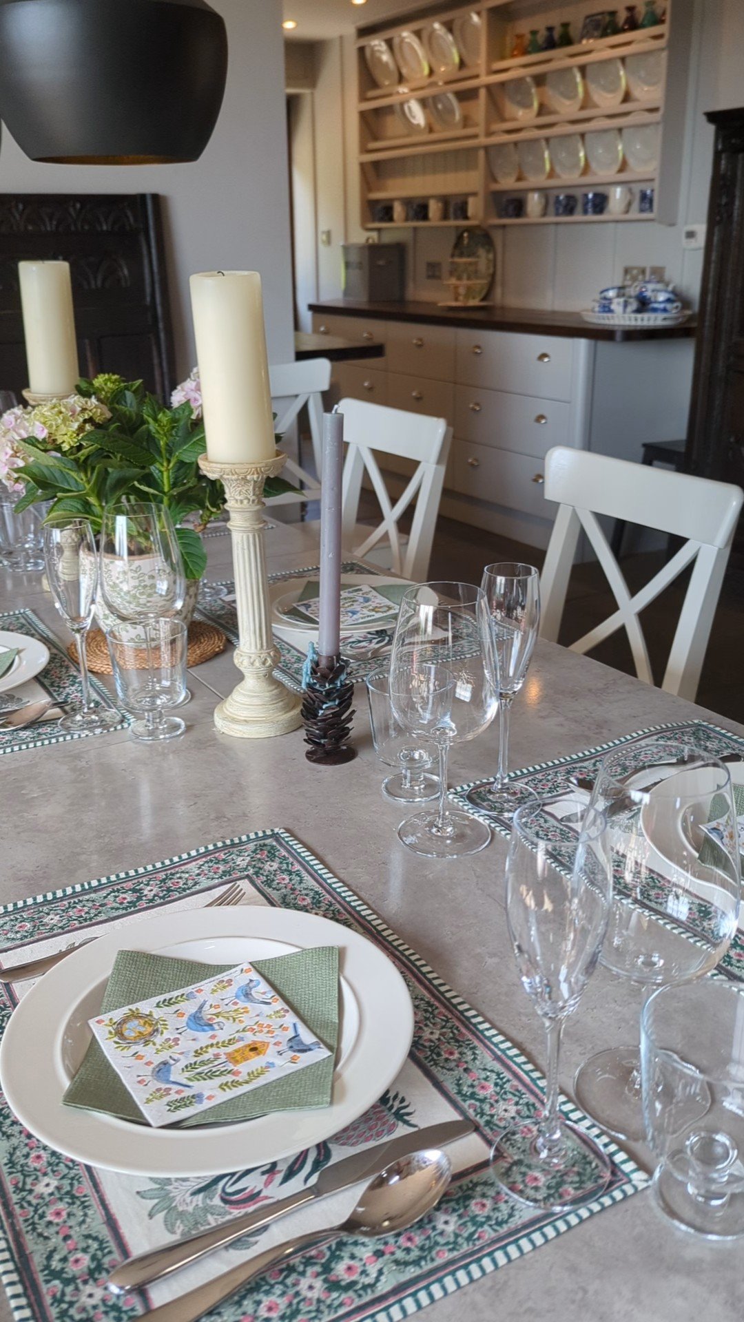 Elegantly set dining table with white plates, floral napkins, wine glasses, and candles in a stylish kitchen. The background has shelves with plates and bowls. A centerpiece with plants adds a decorative touch.