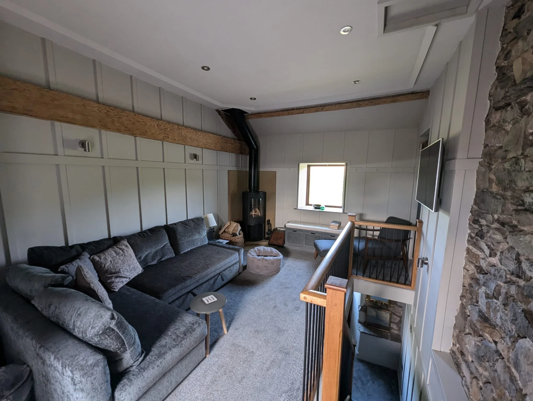 Cozy living room with a gray sectional sofa, wood-burning stove, wooden beams, stone accent wall, and a flat-screen TV mounted on the wall.