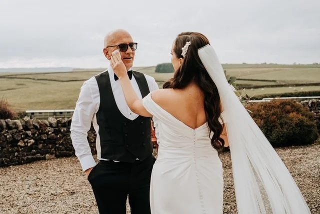 A bride in a white dress and veil wipes the face of a man in a vest and sunglasses, standing in an outdoor rural setting.