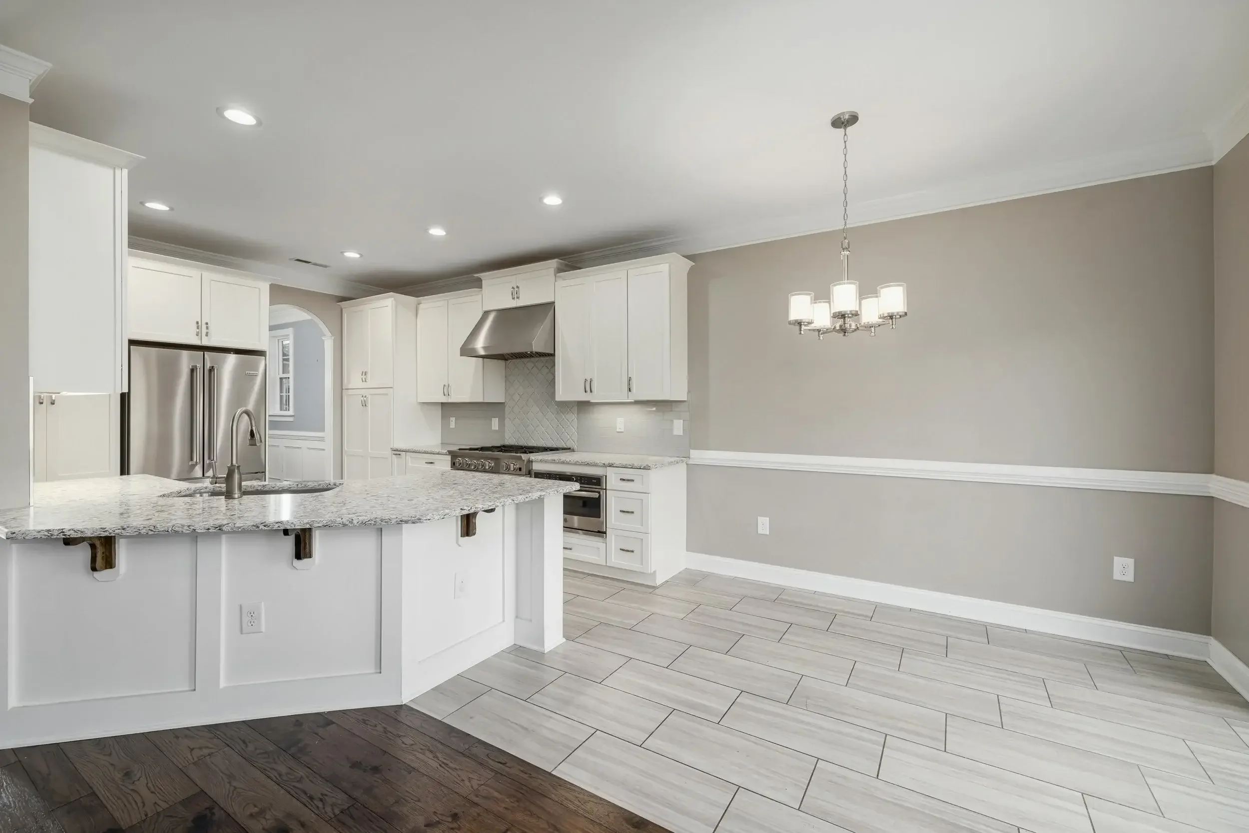 Empty kitchen with white cabinets, stainless steel refrigerator, island with granite countertop, and a dining area with a chandelier.