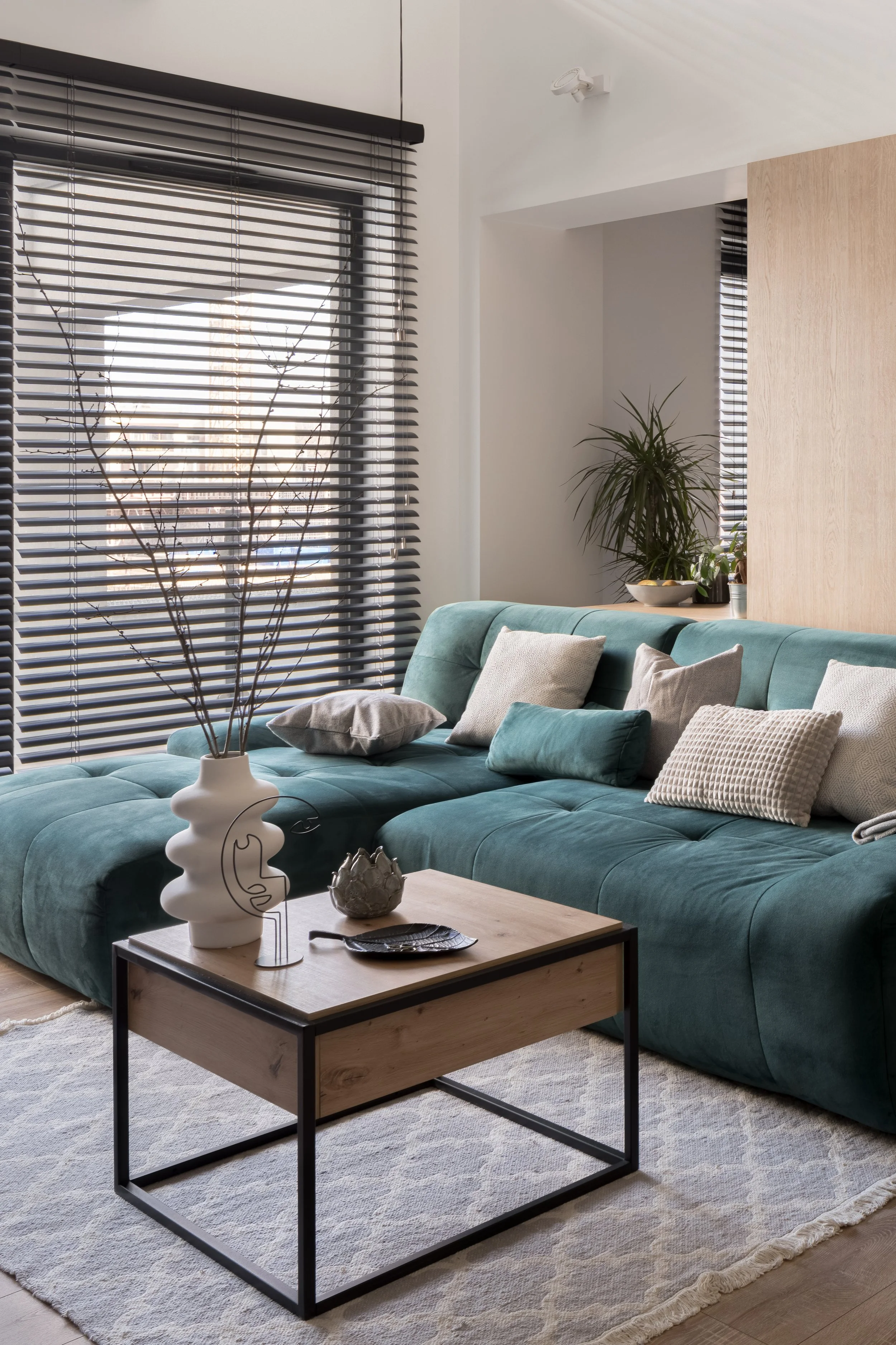 Living room with a teal couch, beige and teal pillows, a wooden coffee table with a white vase and decorative items, a window with black blinds, and plants.