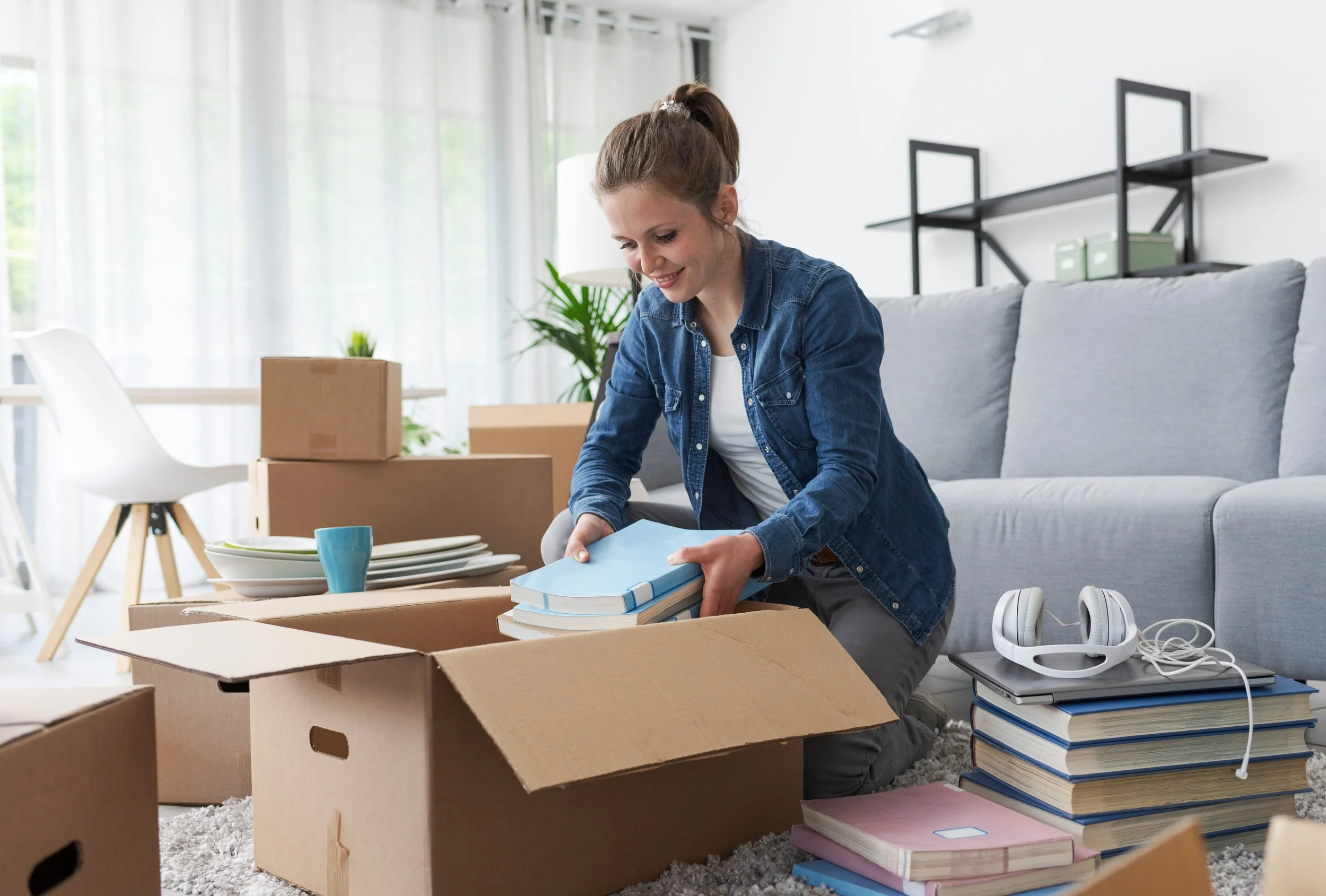 A woman packing books and belongings into moving boxes in a bright living room.