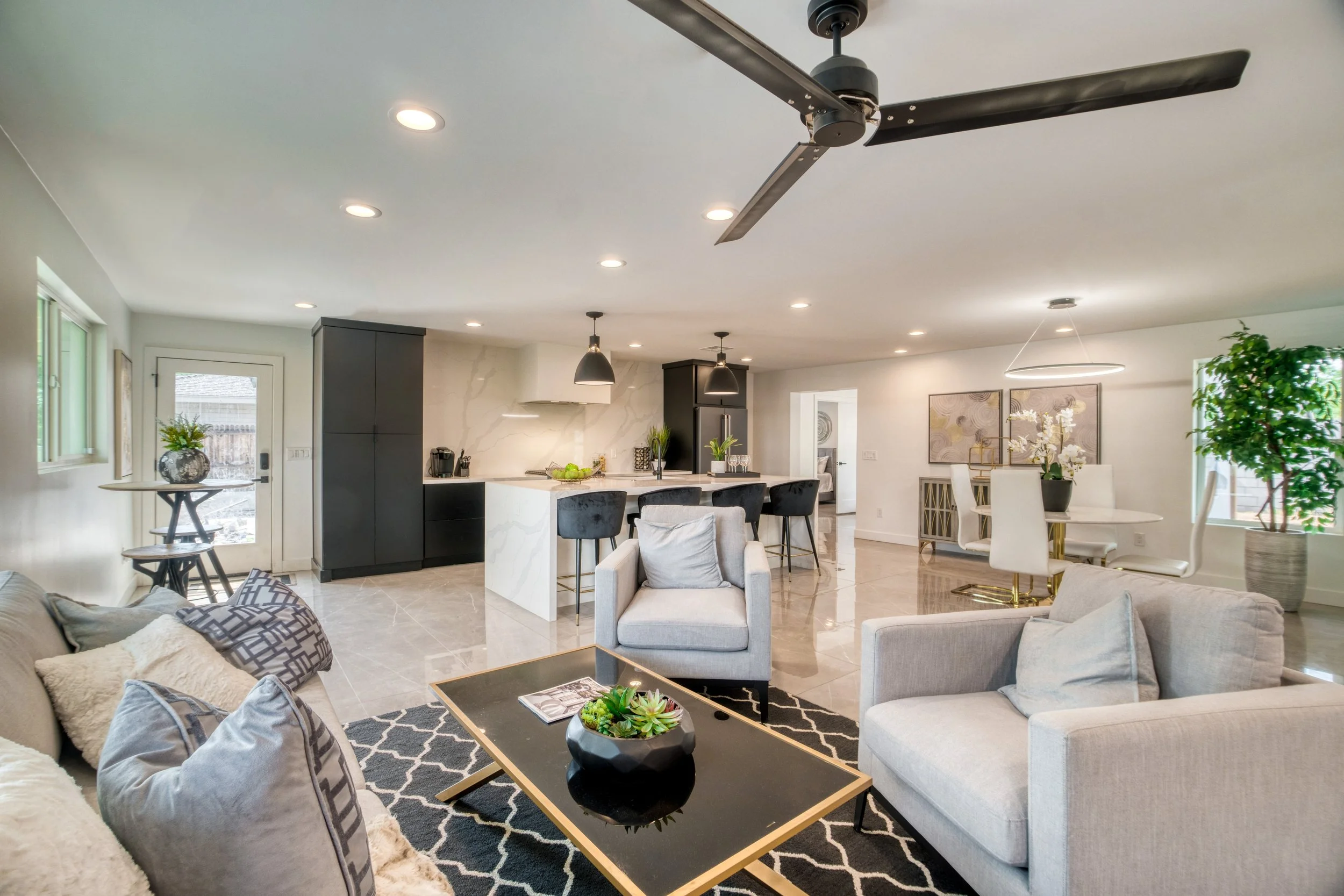 Open-concept living room with beige couches, a black coffee table with a potted plant, a white armchair, modern kitchen with black cabinetry, white marble island, barstools, and dining area with a round table and white chairs, decorated with plants and artwork.