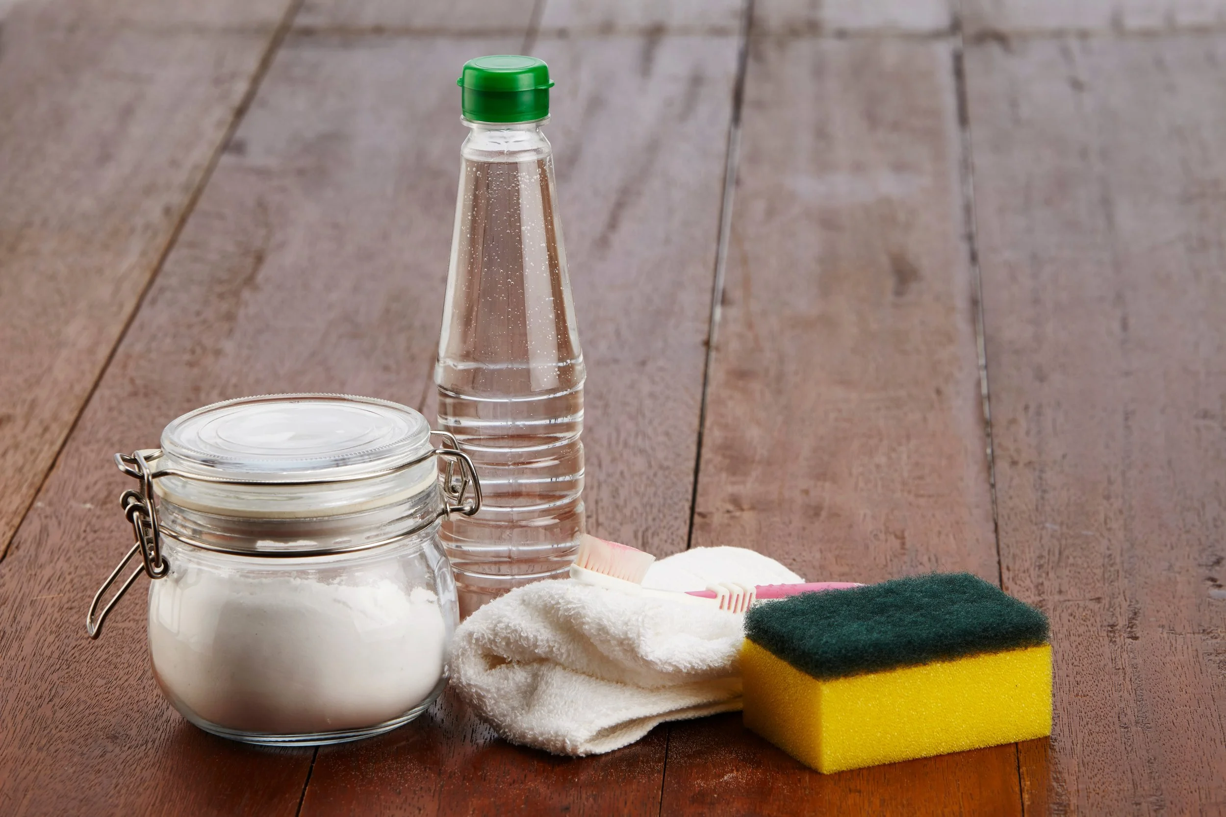 Cleaning supplies on a wooden floor, including a jar of powder, a water bottle, a cleaning cloth, a toothbrush, and a sponge.