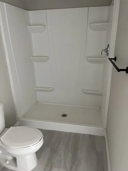 Empty white shower stall with built-in shelves and a drain, adjacent to a toilet in a bathroom.