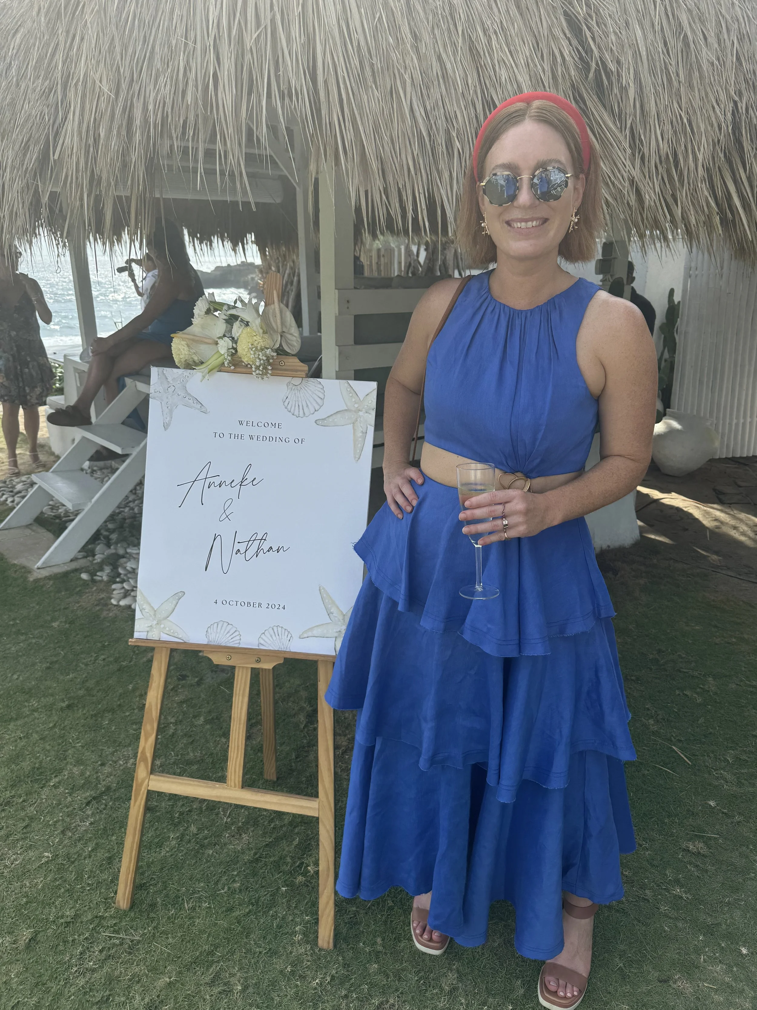 A woman in a blue dress with a red headband and sunglasses holding a drink, standing next to a wedding sign that reads 'Welcome to the wedding of Anike & Nathan, 4 October 2024'. She is smiling at a beachside wedding event, with a thatched roof and other guests in the background.