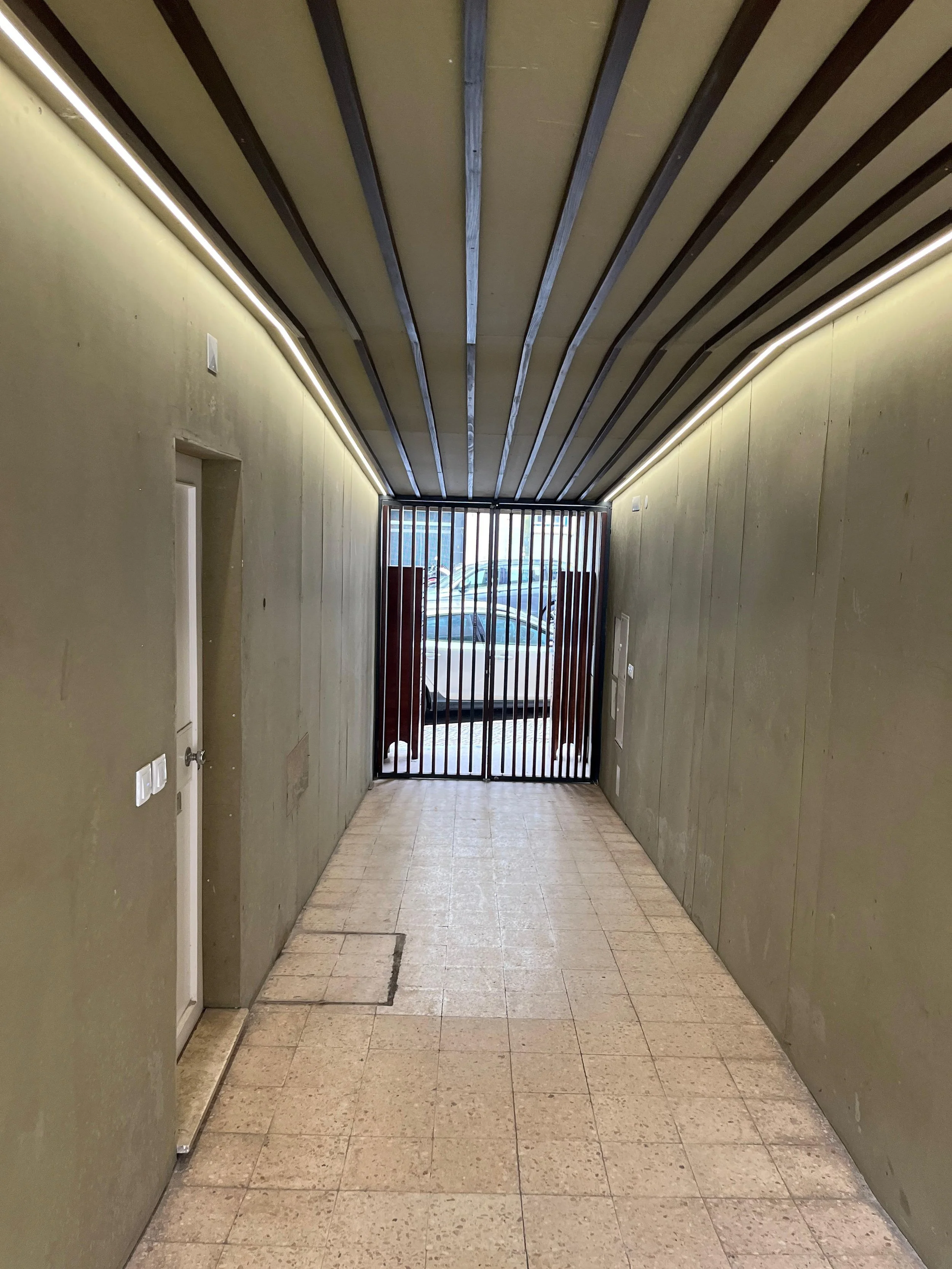 A narrow hallway with olive green walls, a tiled floor, and a ceiling with exposed wooden beams. There is a door on the left side and a decorative wooden slat gate at the end, revealing parked cars outside.