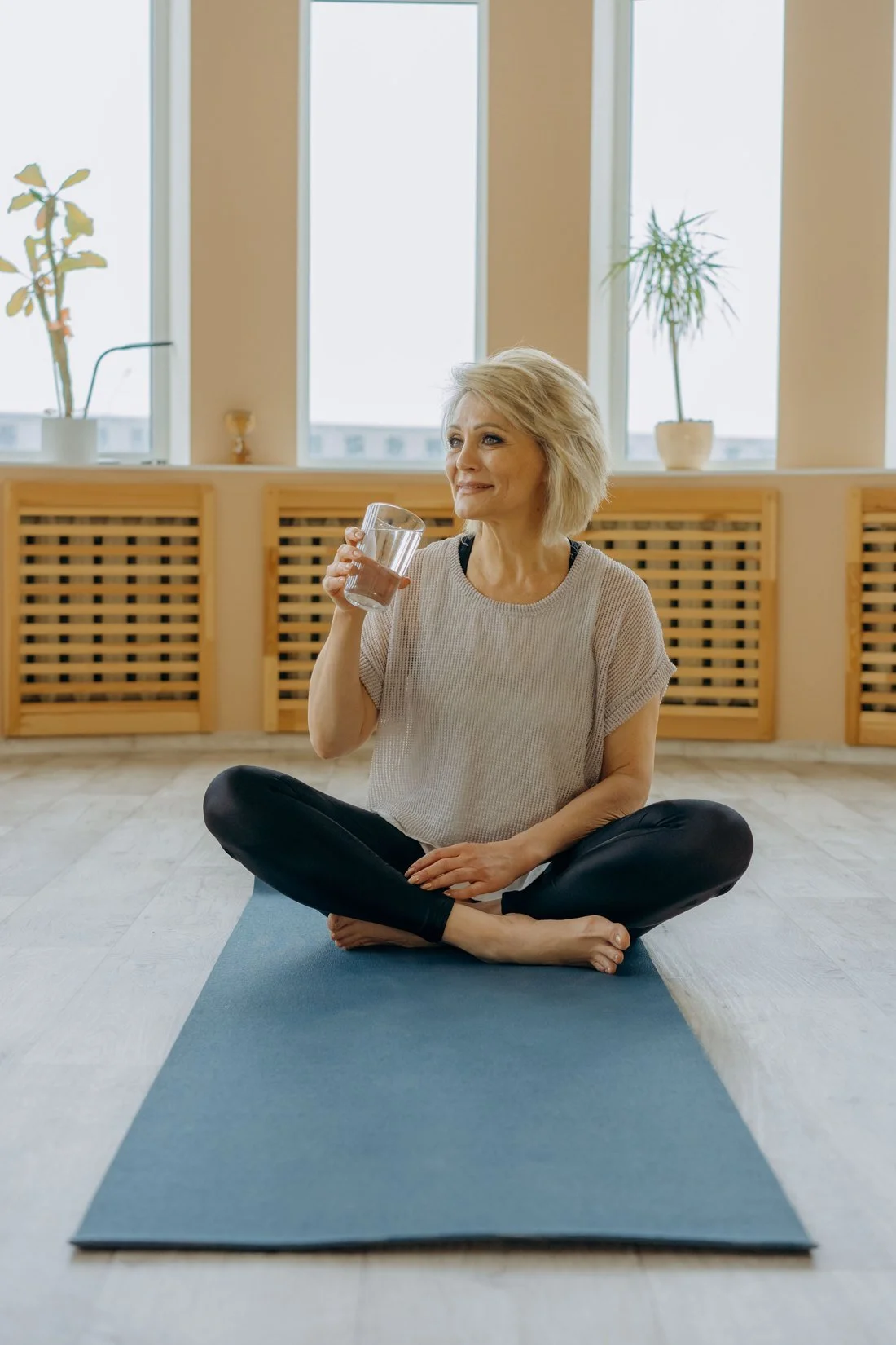 Woman sitting cross-legged on a yoga mat, smiling and drinking a glass of water in a bright room