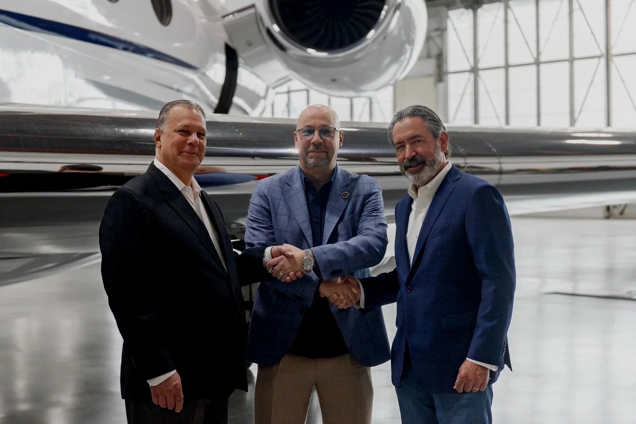 Three men in business attire shaking hands inside an aerospace facility, with a large rocket or spacecraft in the background.