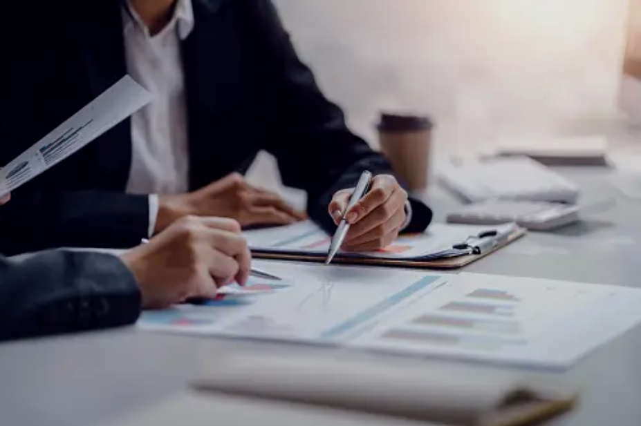 Two people in business attire working on financial charts and documents at a desk in an office.