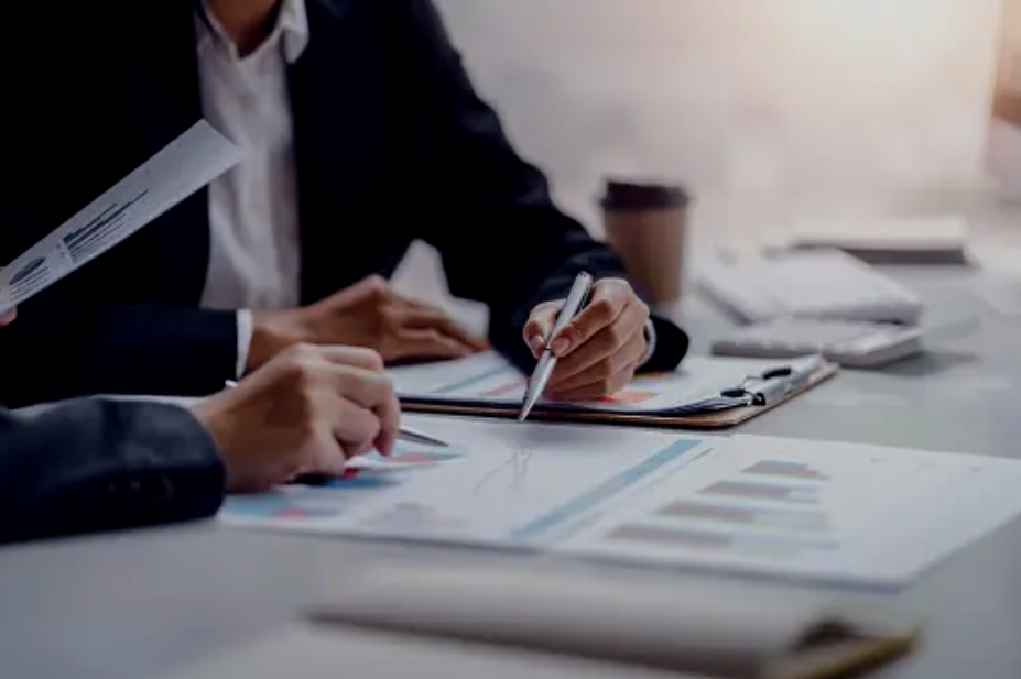 Two people in business attire working on financial charts and documents at a desk in an office.