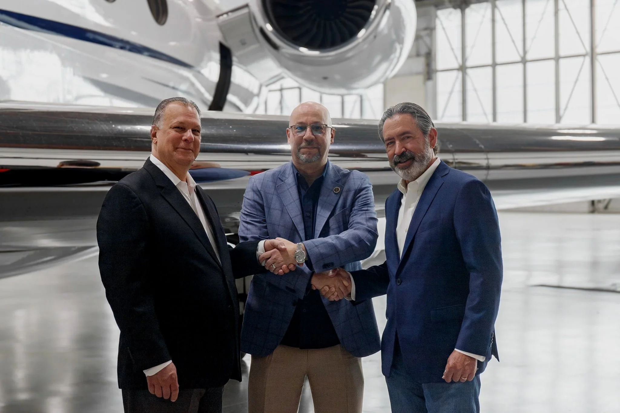 Three men in business attire shaking hands inside an aerospace facility, with a large rocket or spacecraft in the background.