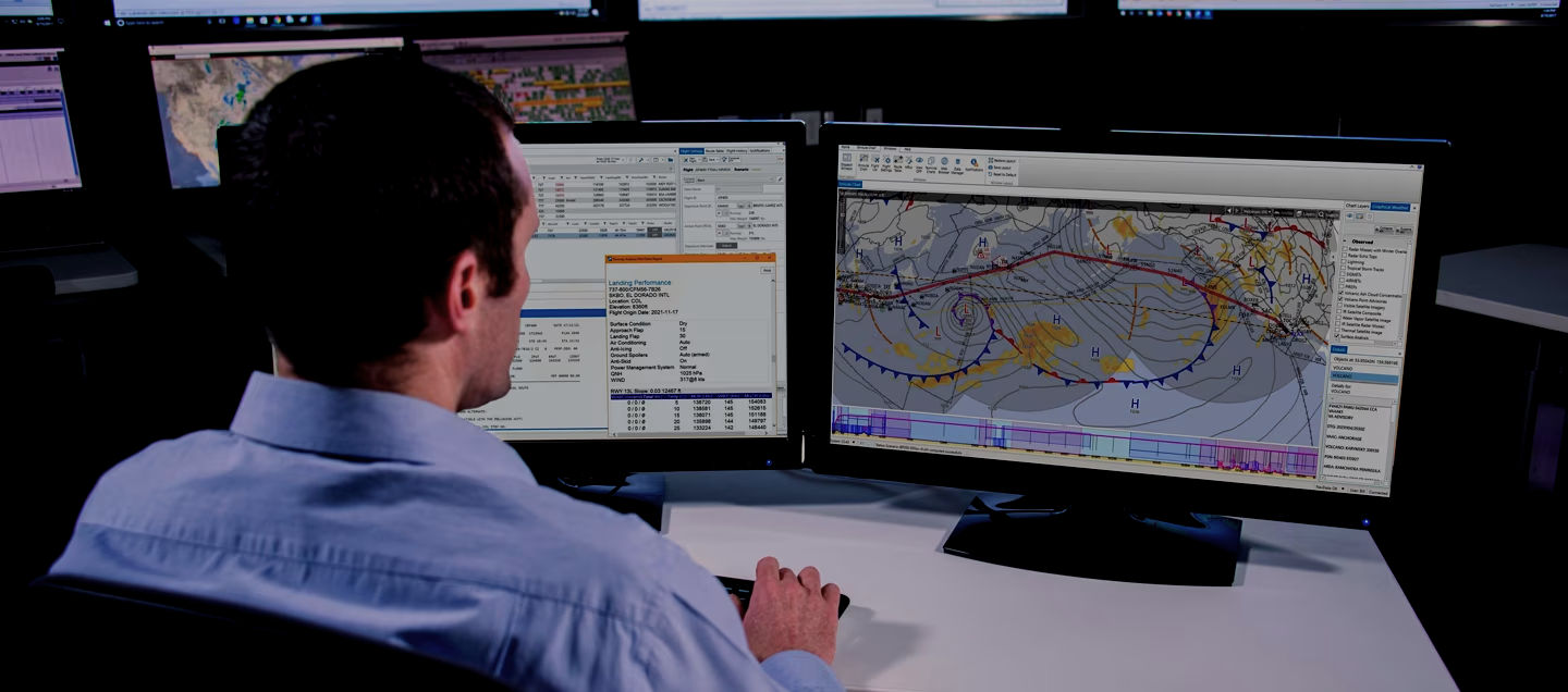 A man working at a desk with two large computer monitors displaying weather and flight data, maps, and charts.