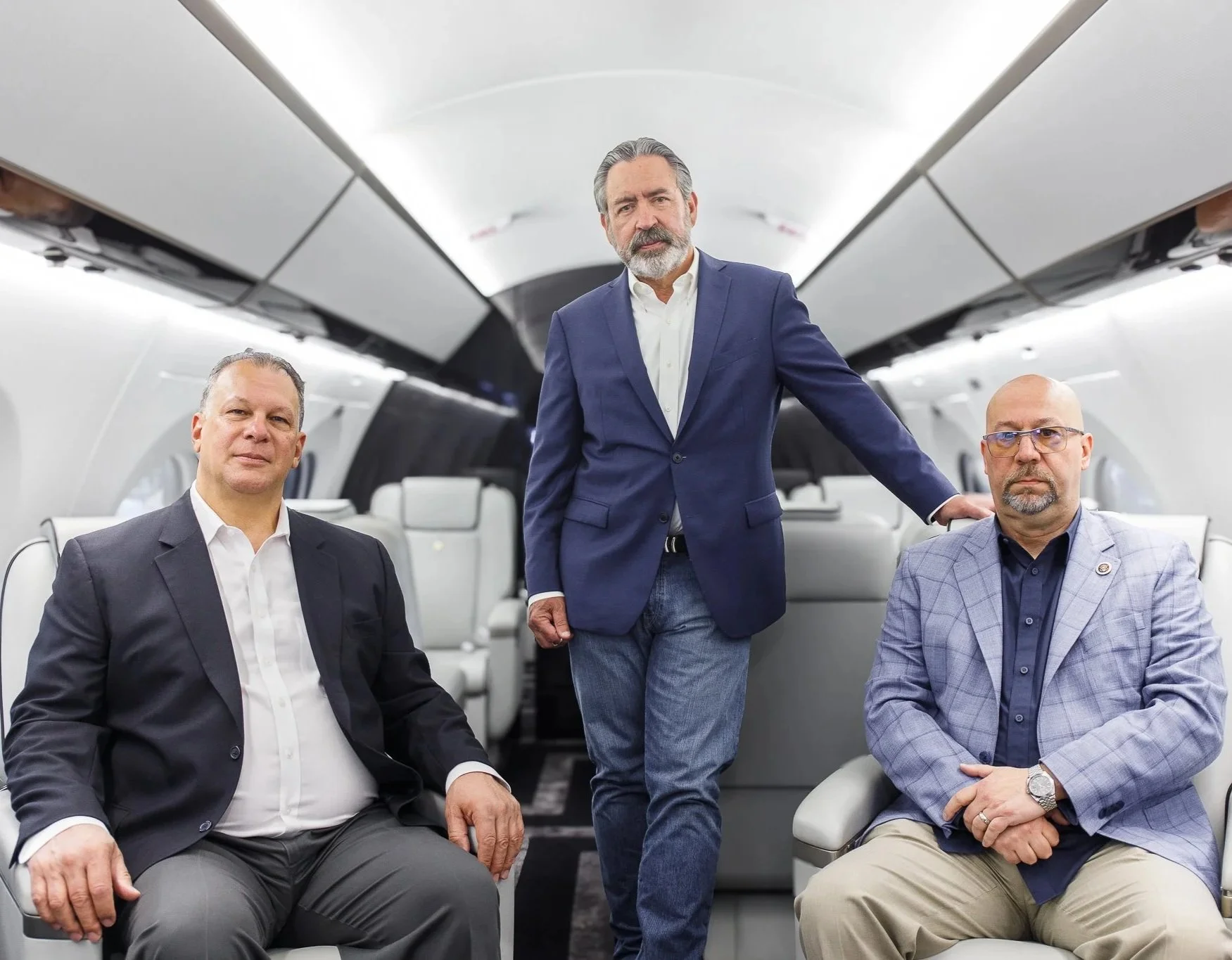 Three middle-aged men in suits inside a luxurious airplane, with two seated and one standing. The man in the center is standing, wearing a dark blue blazer and jeans. The two men seated are wearing light-colored blazers and shirts.