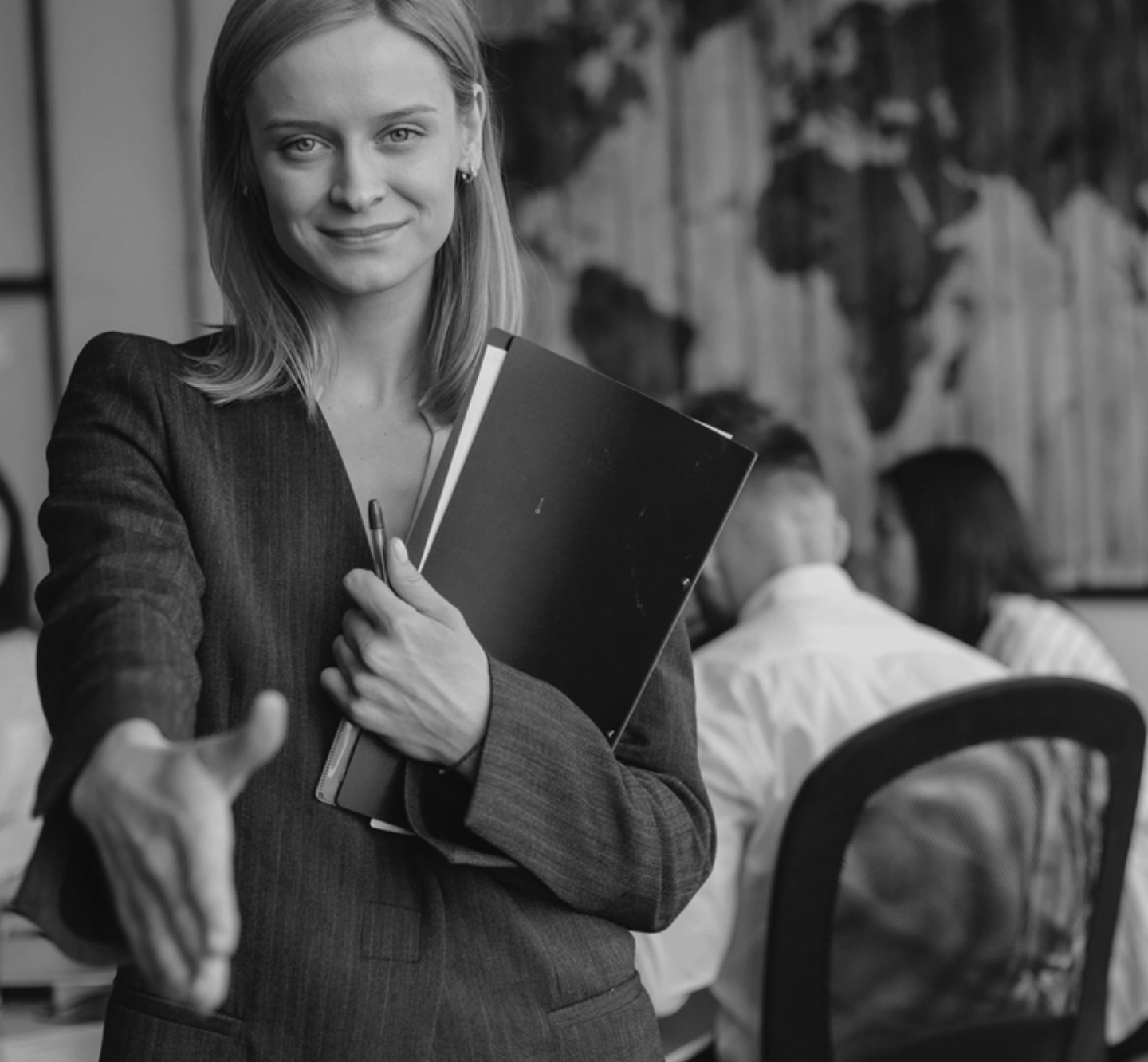 A woman in a business suit holding a folder and pen, reaching out for a handshake in an office setting.