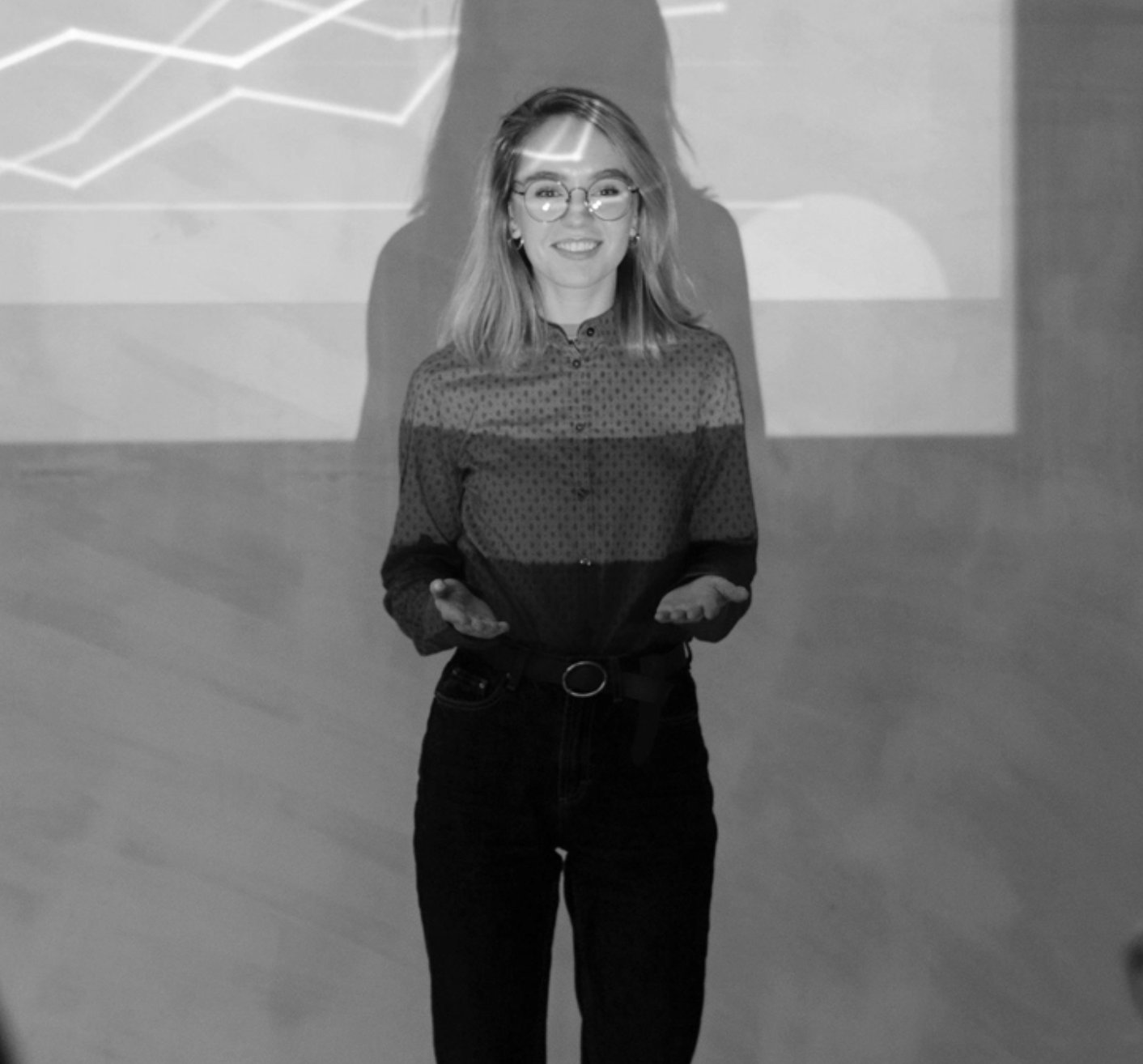 A woman with glasses and shoulder-length hair, smiling and gesturing with her hands, standing in front of a projected presentation with line graphs.