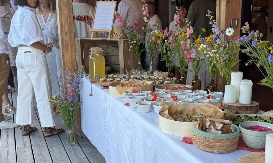 Buffet de nourriture avec fleurs décoratives dans des vases en verre et en céramique, sur une table blanche en bois, lors d'un événement en plein air.