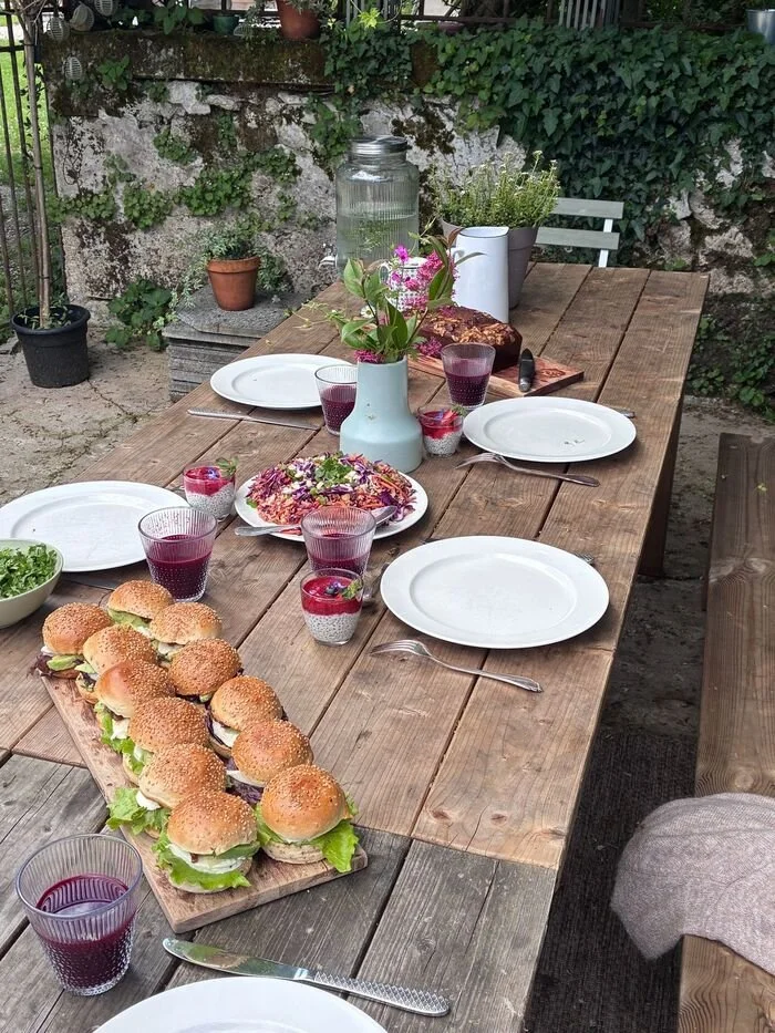 Table d'extérieur prête pour un repas, avec des burgers végétariens, des verres de jus de betterave, une tarte, une plante en vase, un grand pichet en verre avec de l'eau, et quelques assiettes et couverts