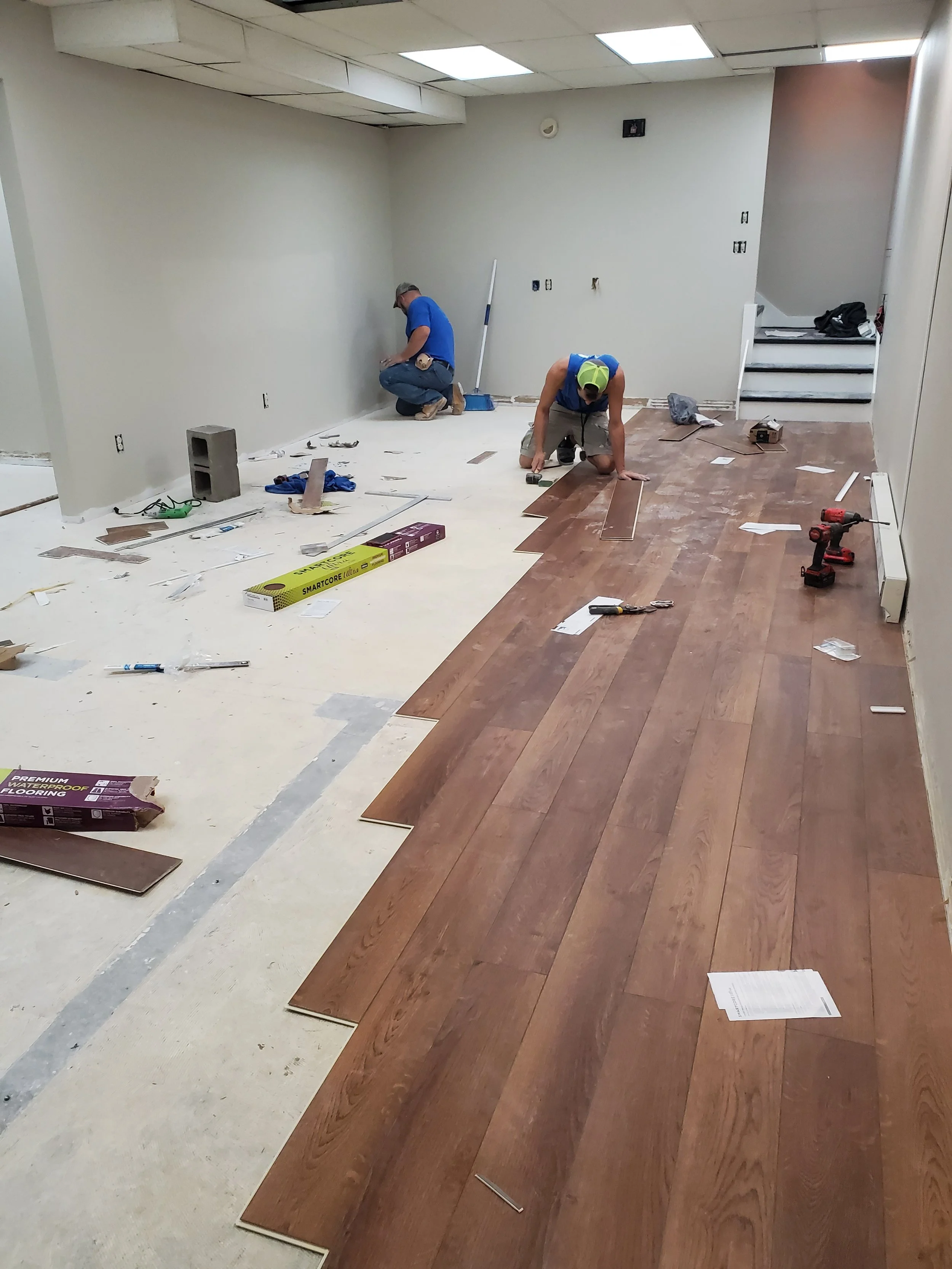 Two workers installing hardwood flooring in a room with white walls and ceiling. Construction tools, materials, and scattered papers are visible on the floor.