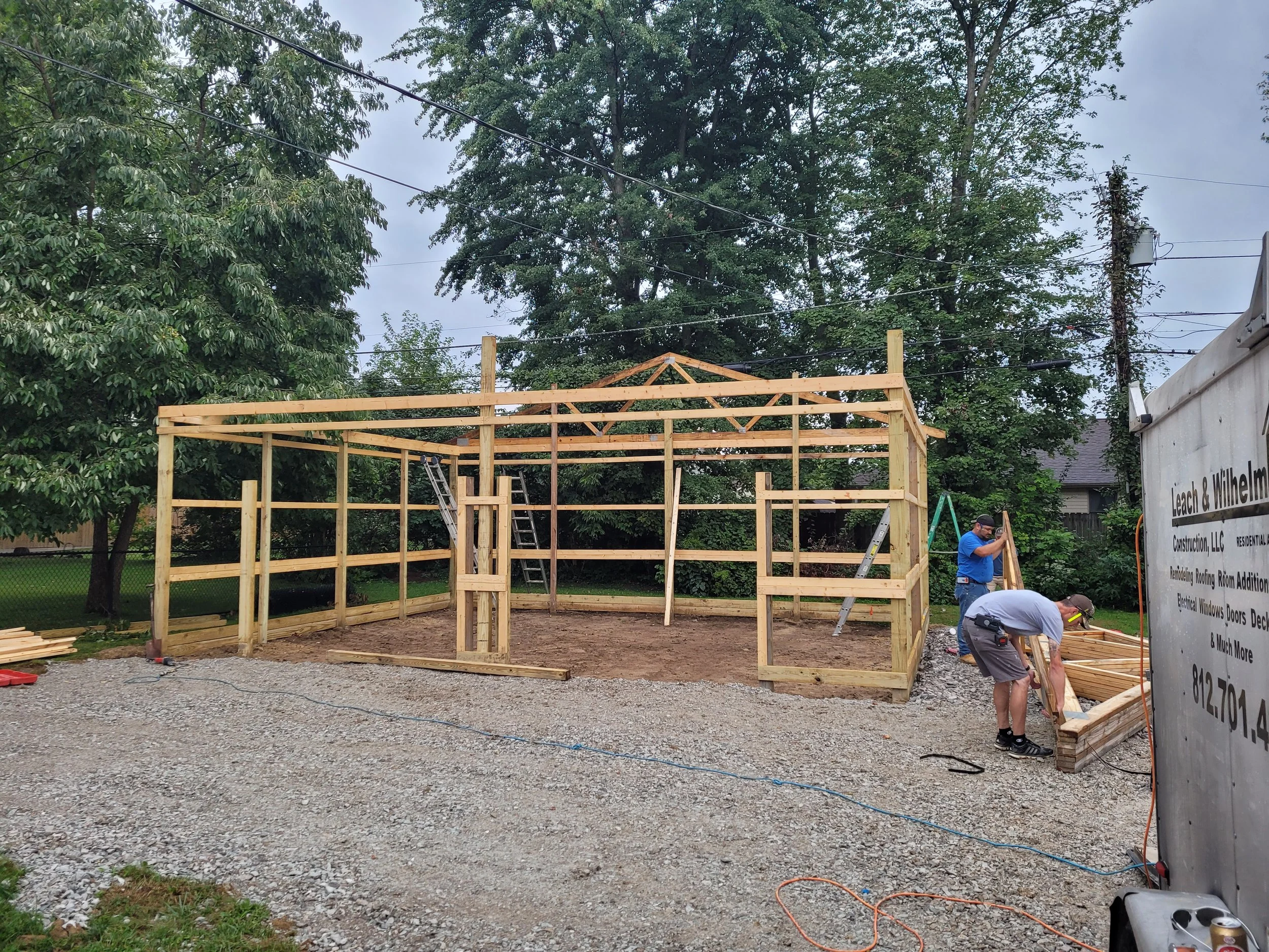 Two construction workers building a wooden frame structure in a backyard on a gravel surface, with green trees and a neighboring house in the background.