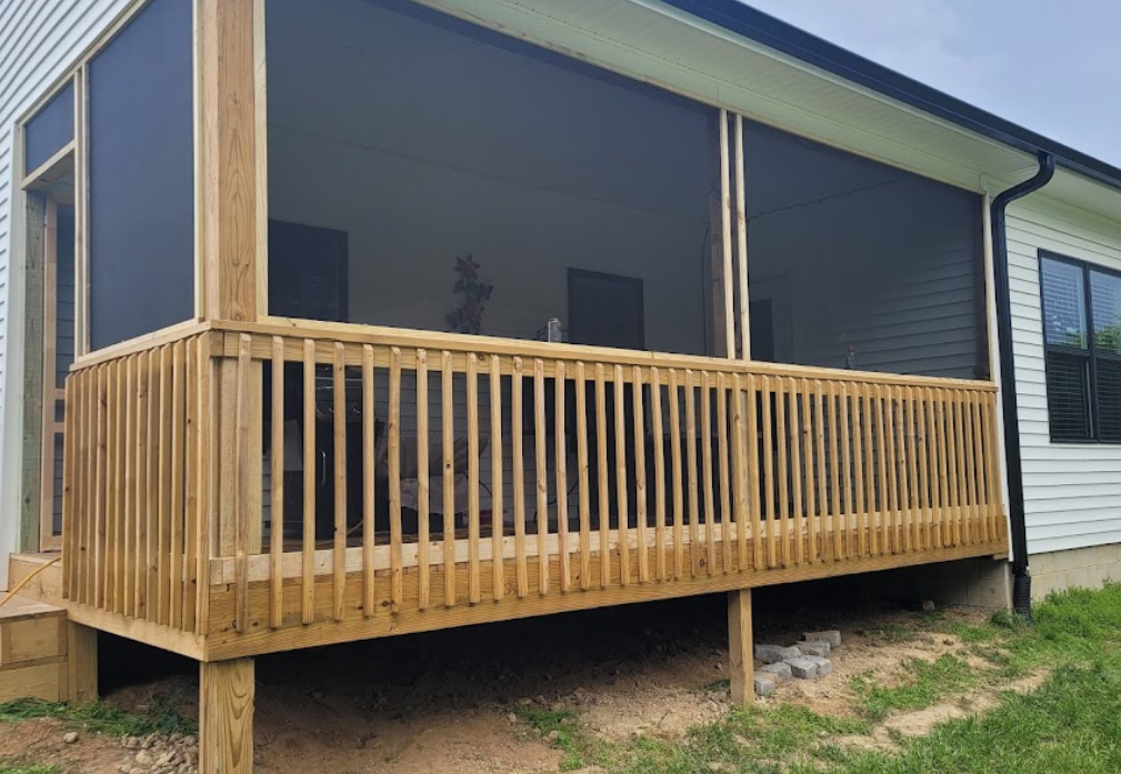Newly built screened-in wooden porch attached to a house with white siding, supported by wooden beams, with a concrete foundation and grassy yard underneath.