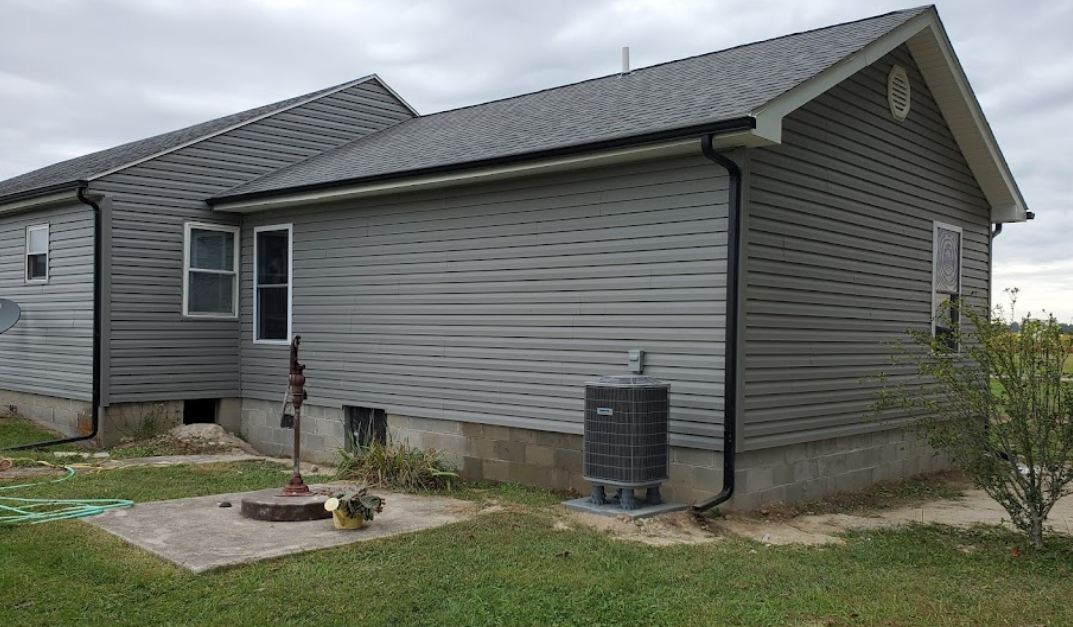 Side view of a gray house with vinyl siding, black gutters, an air conditioning unit, and a small tree in the yard.