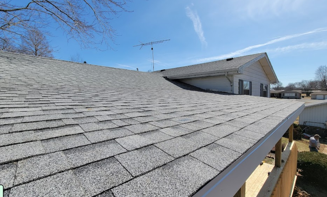 Close-up view of a gray asphalt shingle roof on a house with a clear blue sky and some trees in the background.