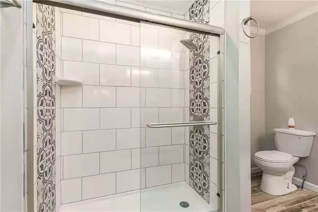 Bathroom with a walk-in shower featuring white subway tiles and decorative gray tile accents, a glass shower door, and a toilet with a bottle of personal care product on top.