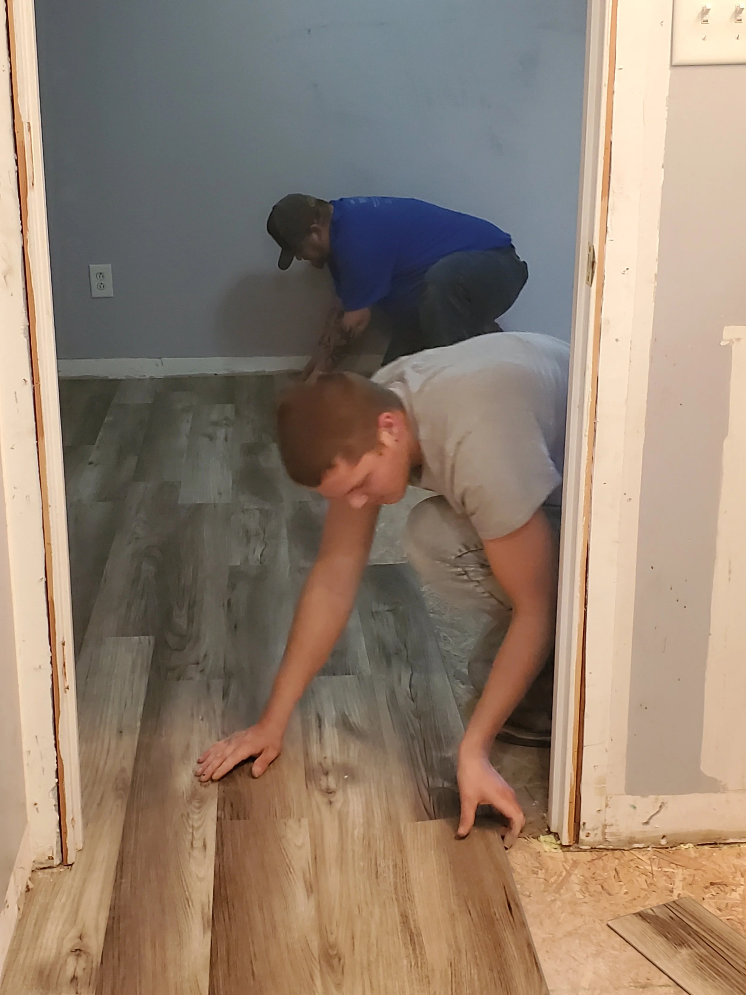 Two men installing hardwood flooring in a room, one kneeling and one bending over, working together.