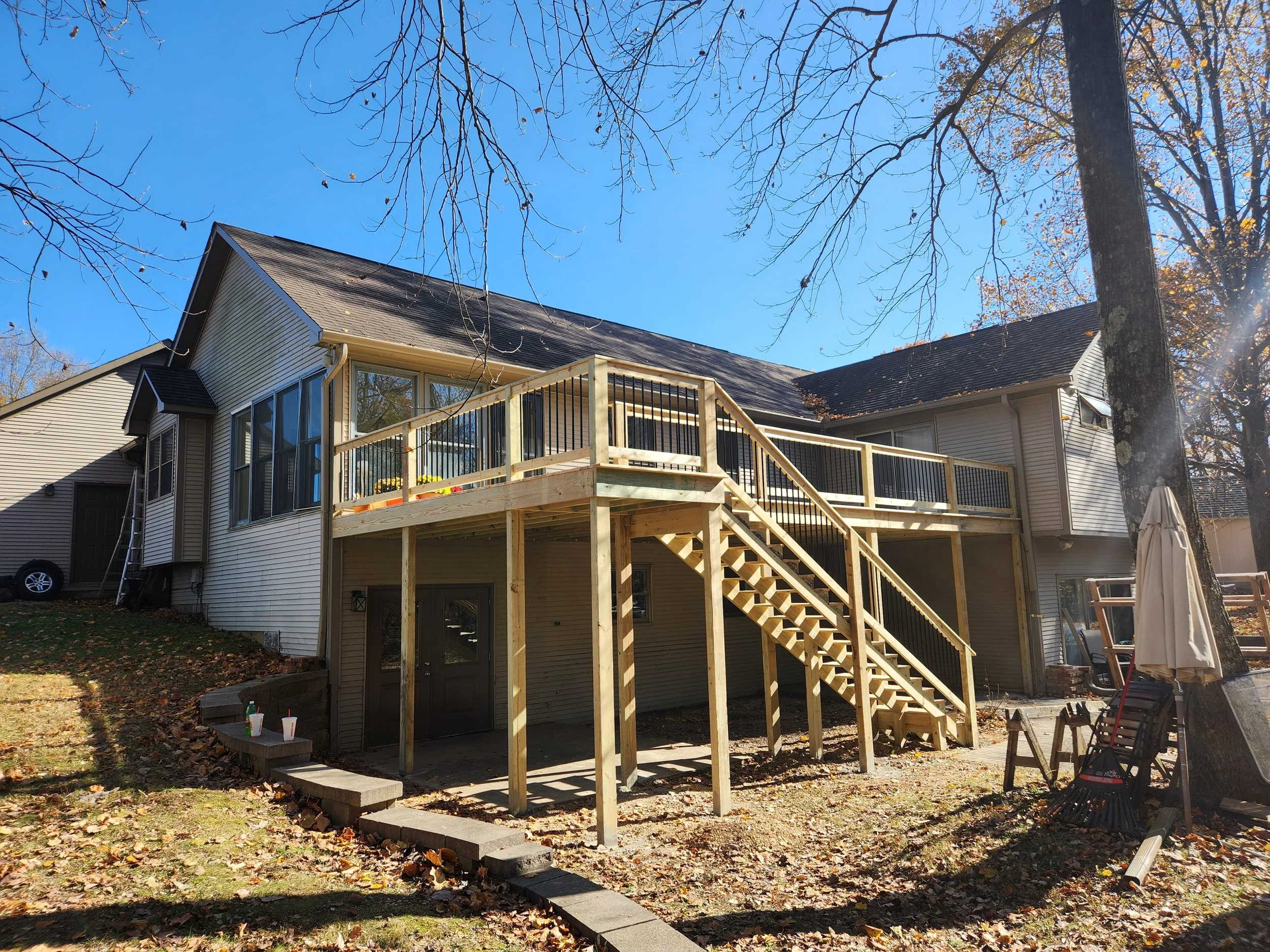 Residential house with an elevated wooden deck under construction, with stairs leading to the deck, and various tools and materials in the yard.