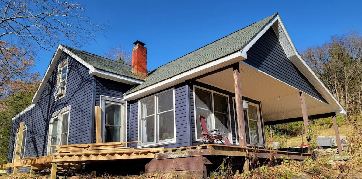 A two-story house with blue vinyl siding, a brick chimney, a covered porch with a rocking chair, and a partially constructed wooden deck on a raised foundation. The house is surrounded by trees and has a clear blue sky in the background.