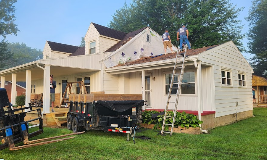 Construction workers repairing a roof on a white house during daytime, with tools and equipment in the yard, and a trailer nearby.