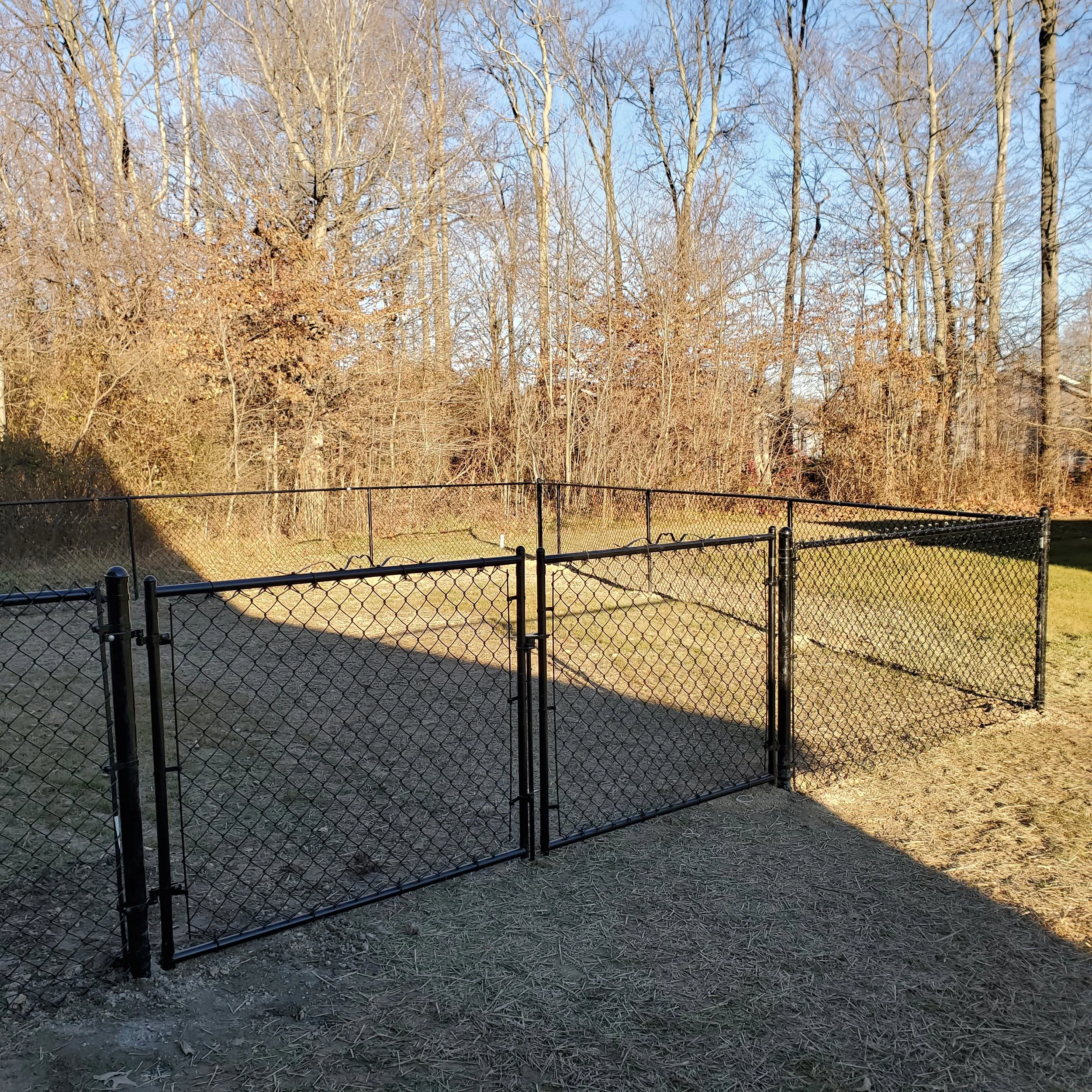 A black chain-link fence yard enclosure in a backyard with dry grass and leafless trees in the background, under a clear blue sky.