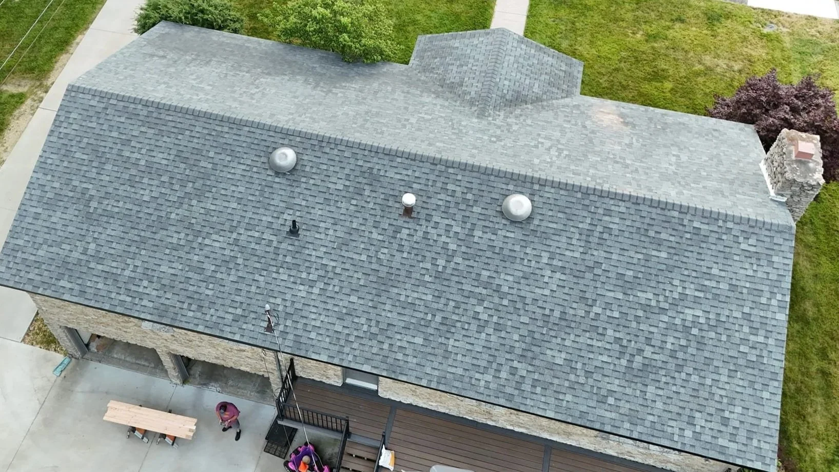 Aerial view of a house with a gray shingle roof, surrounded by a grassy yard, sidewalk, and sidewalk with some people and outdoor furniture.