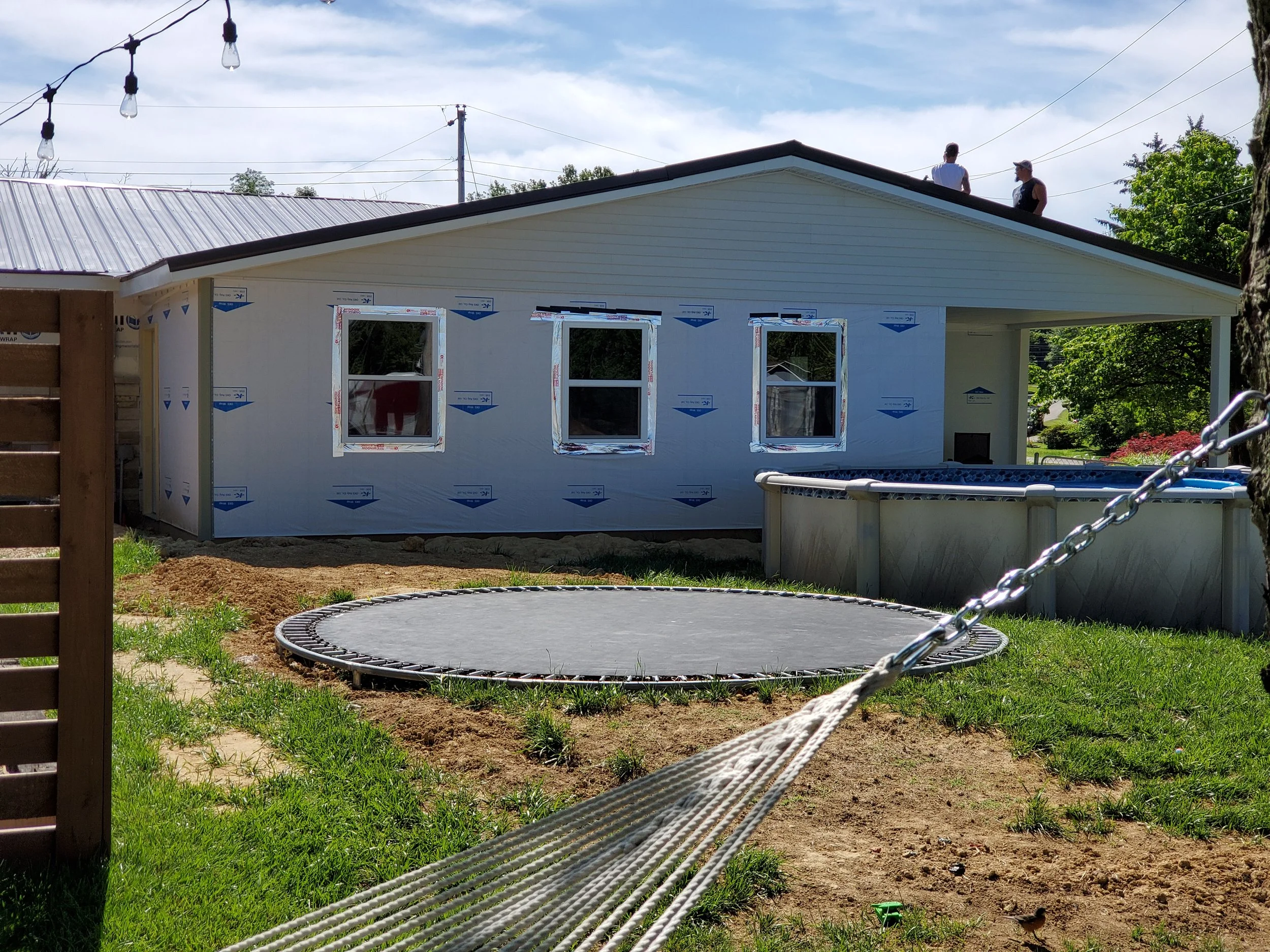 House under construction with windows, a trampoline, a pool, and two people on the roof, with outdoor string lights and trees in the background.