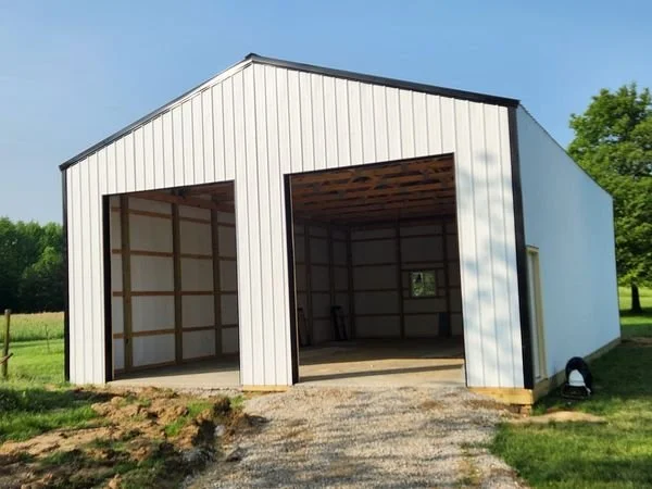 A large white metal building with two open garage bays on a grassy lot with trees in the background.