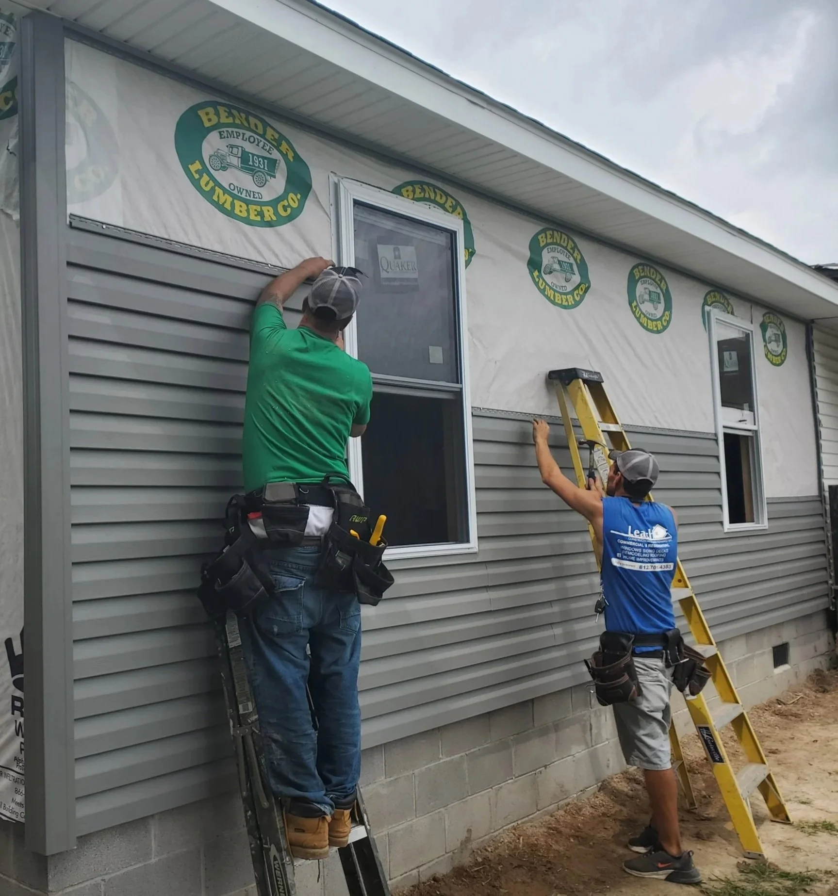 Two workers installing or repairing windows on a house. One is on a ladder reaching the window, while the other stands on the ground holding a ladder in place. The house has siding and logo stickers that say 'Bender Lumber Co.'