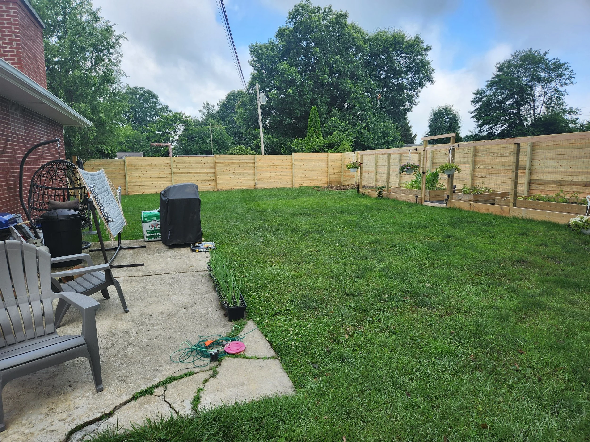 Backyard with a concrete patio and a grassy lawn, enclosed by a wooden fence. There are outdoor chairs, a grill, and gardening tools on the patio, with some potted plants and hanging flower baskets along the fence.