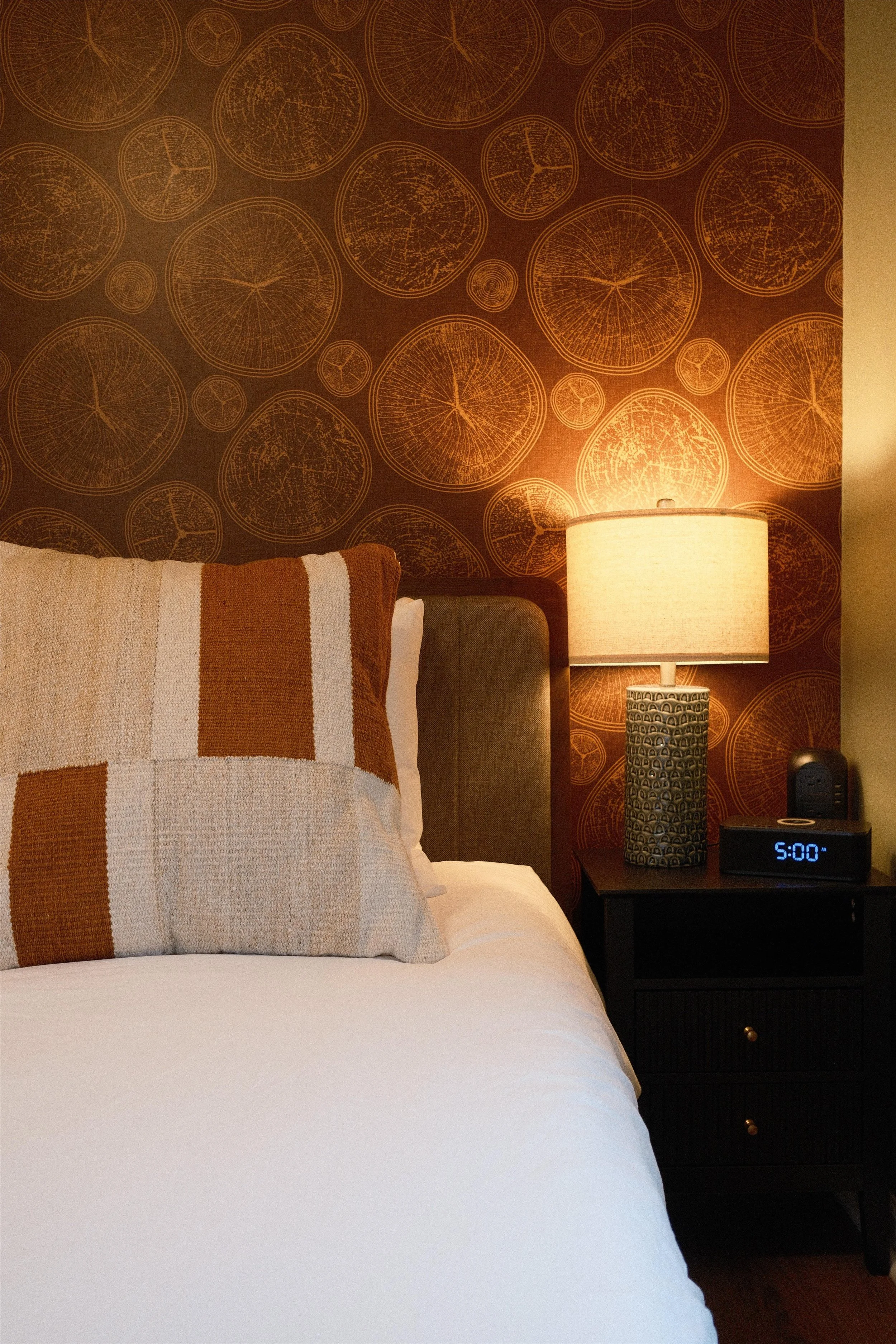 A bedroom bedside table with a modern lamp, digital clock showing 5:00, and a speaker against a wall with brown, circular patterned wallpaper.