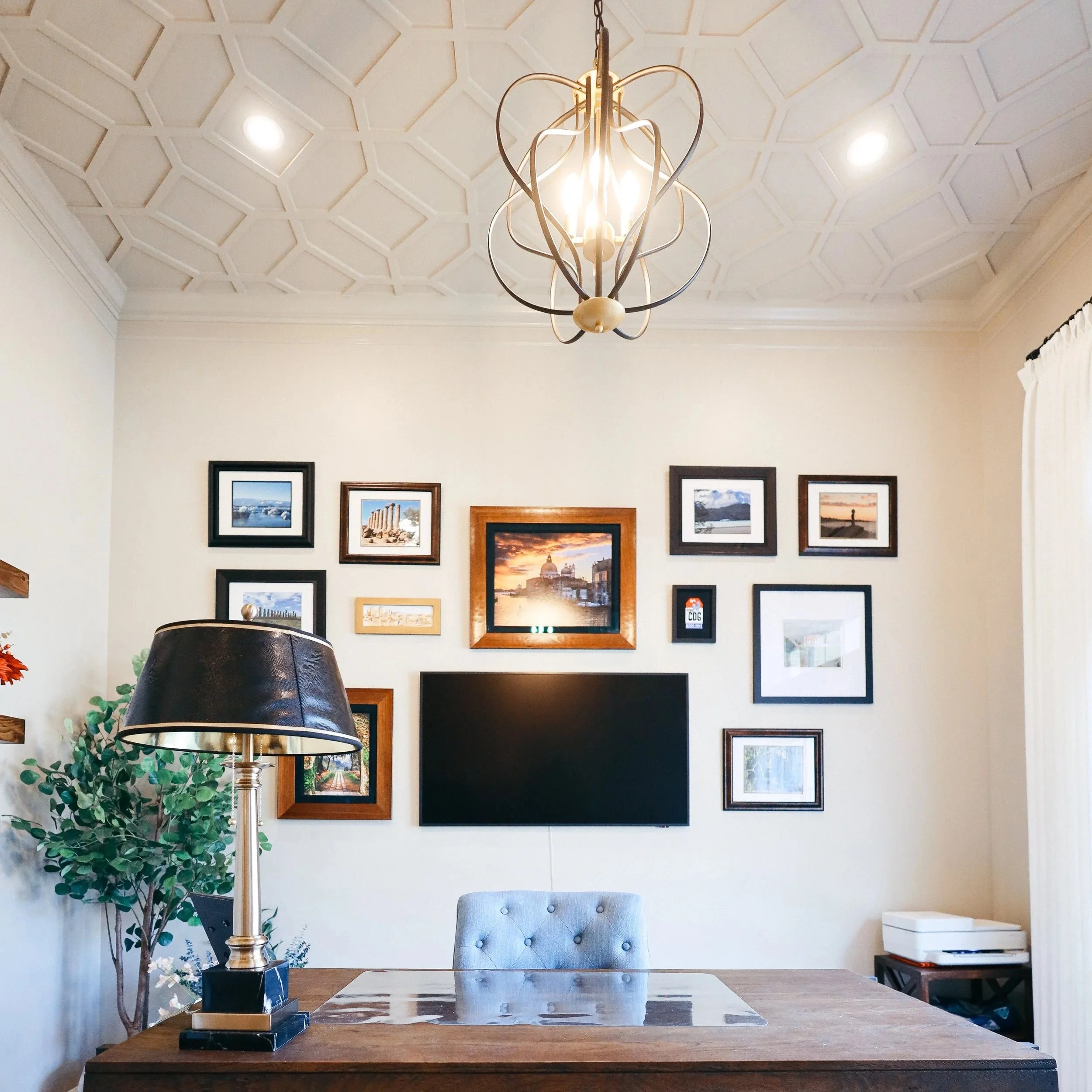 A home office with a wooden desk, grey tufted chair, a large patterned rug, a black table lamp, a gallery wall with framed photos and artwork, a wall-mounted flat-screen TV, a potted plant, and a chandelier hanging from the ceiling.