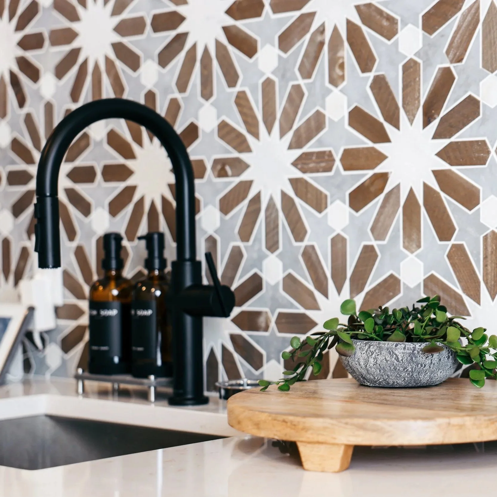 Modern kitchen with black faucet, amber soap dispensers, a gray bowl with a green plant, and geometric-patterned brown and white tile backsplash.