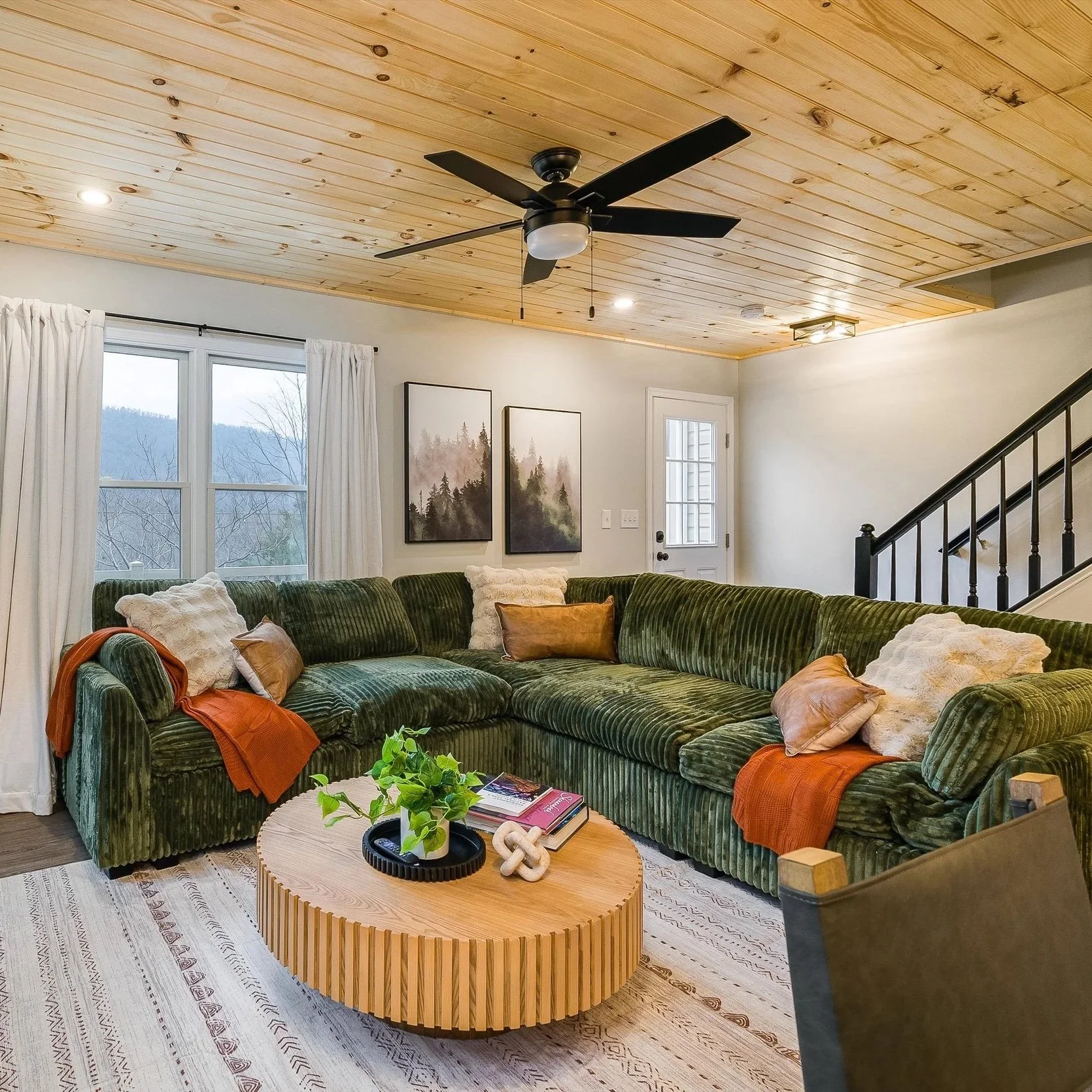 Living room with a large green velvet sectional sofa, wooden coffee table, and framed forest landscape artwork on the wall. Window with white curtains, ceiling fan, staircase railing, and door in the background.