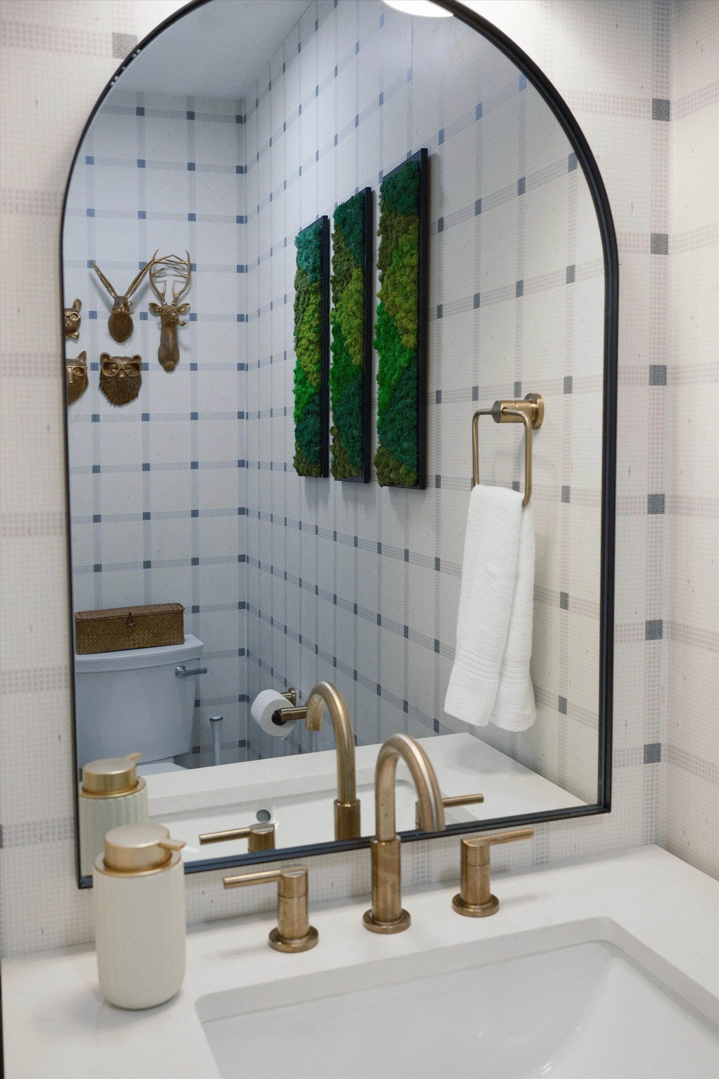 A bathroom mirror reflecting a toilet, a gold faucet, soap dispenser, and wall decor including greenery, mounted animal heads, and masks.