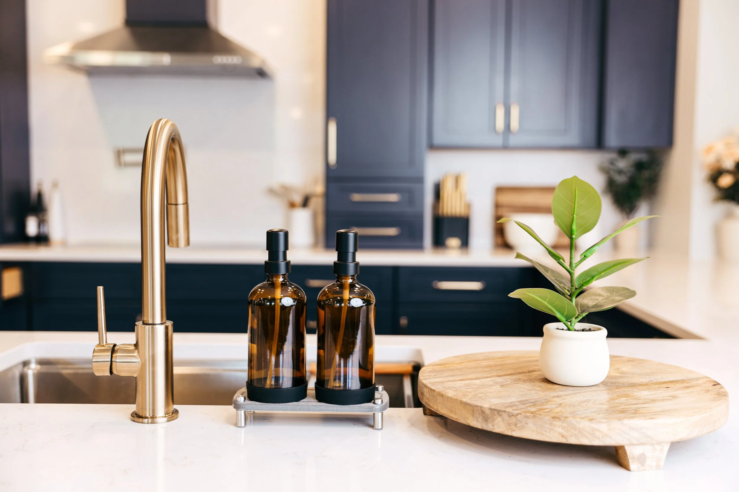 Kitchen countertop with a gold faucet, two amber glass bottles with black caps, a round wooden tray, and a potted plant in a white pot, with dark blue cabinets in the background.