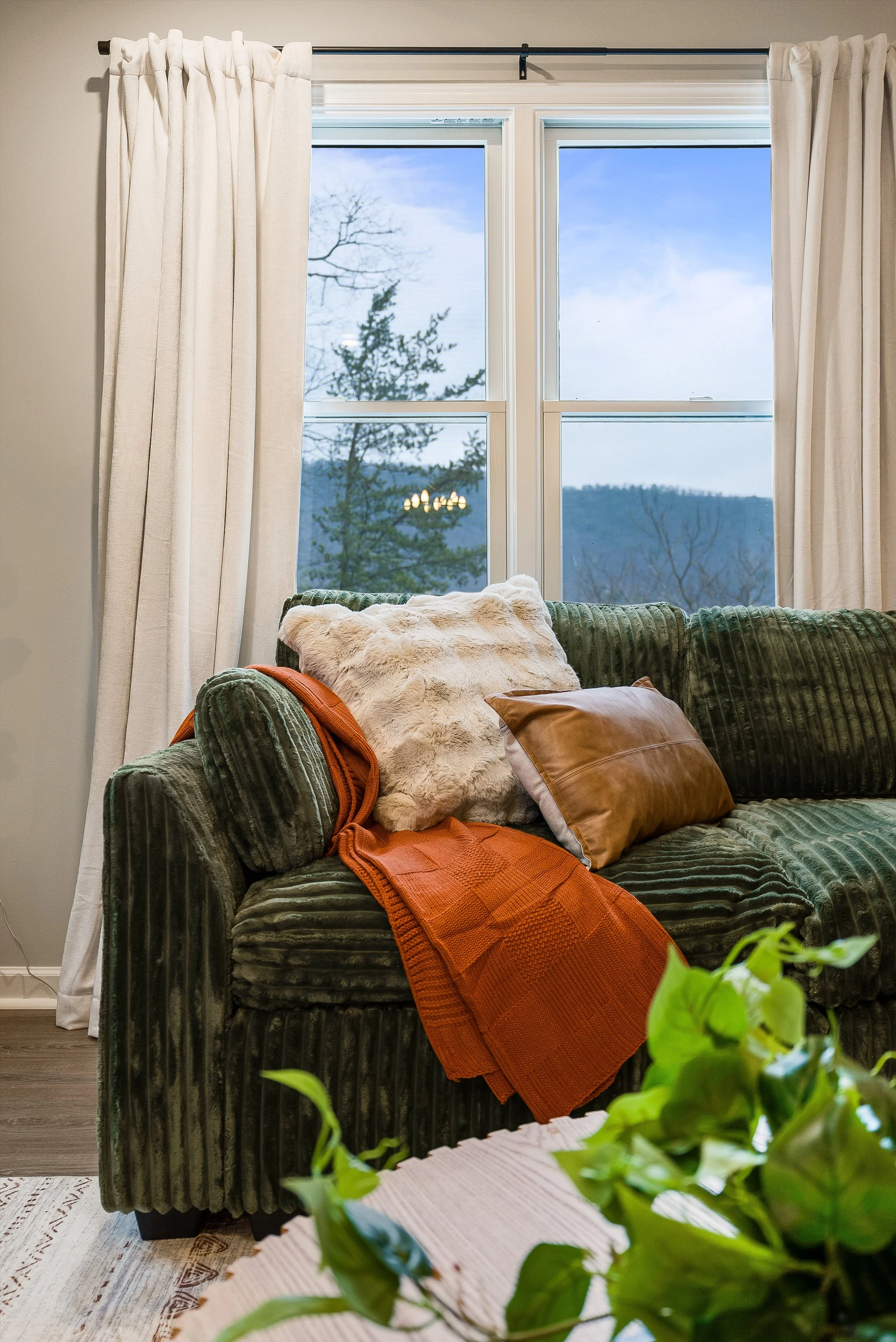 A living room with a green velvet sofa, decorated with a brown leather pillow, a shearling pillow, and an orange blanket. Behind the sofa, a large window with white curtains looks out onto a mountainous landscape with trees. Green plant leaves are visible in the foreground.