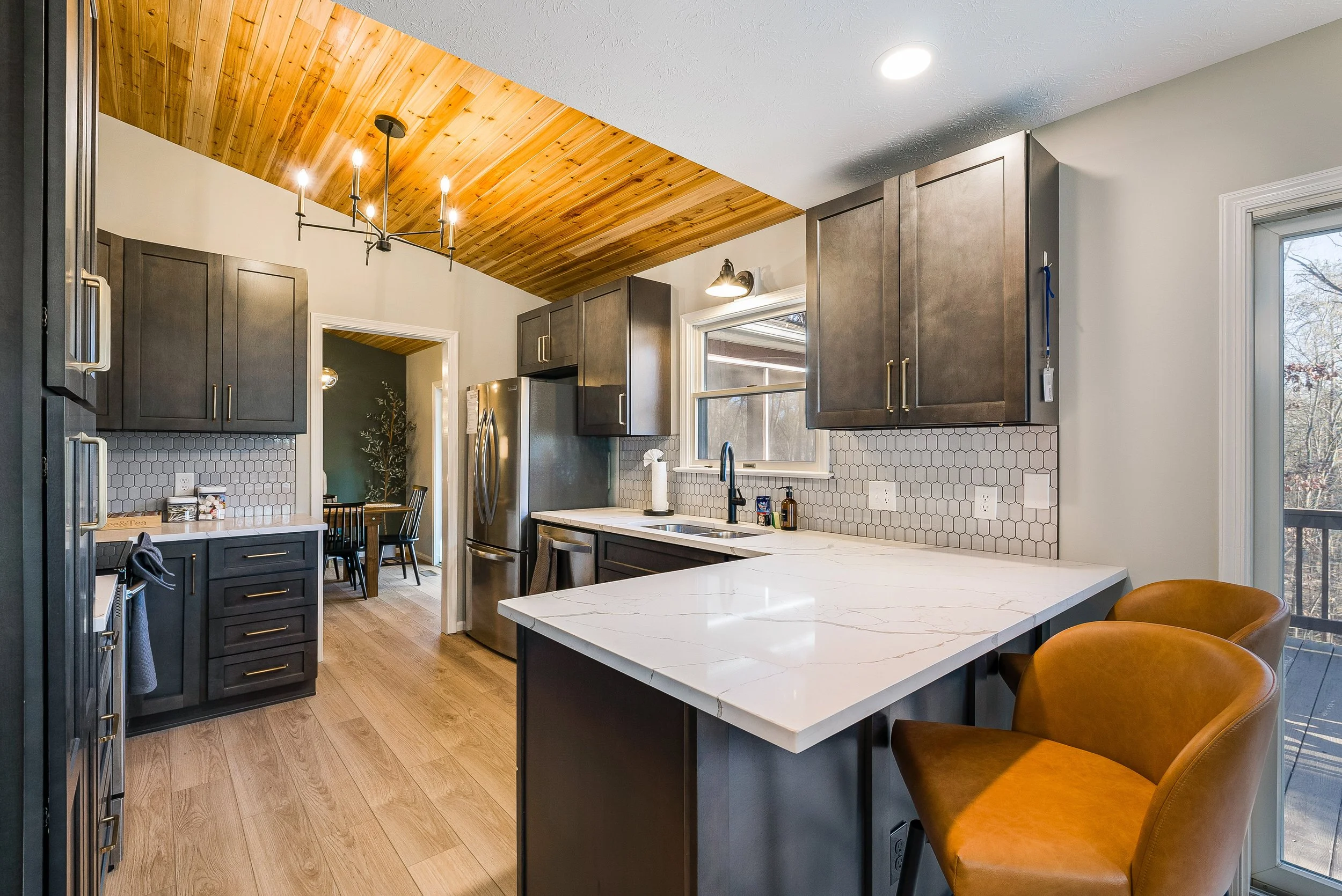 Modern kitchen with dark cabinets, white marble countertop, wood ceiling, stainless steel refrigerator, hexagonal tile backsplash, and two orange chairs by a sliding glass door.