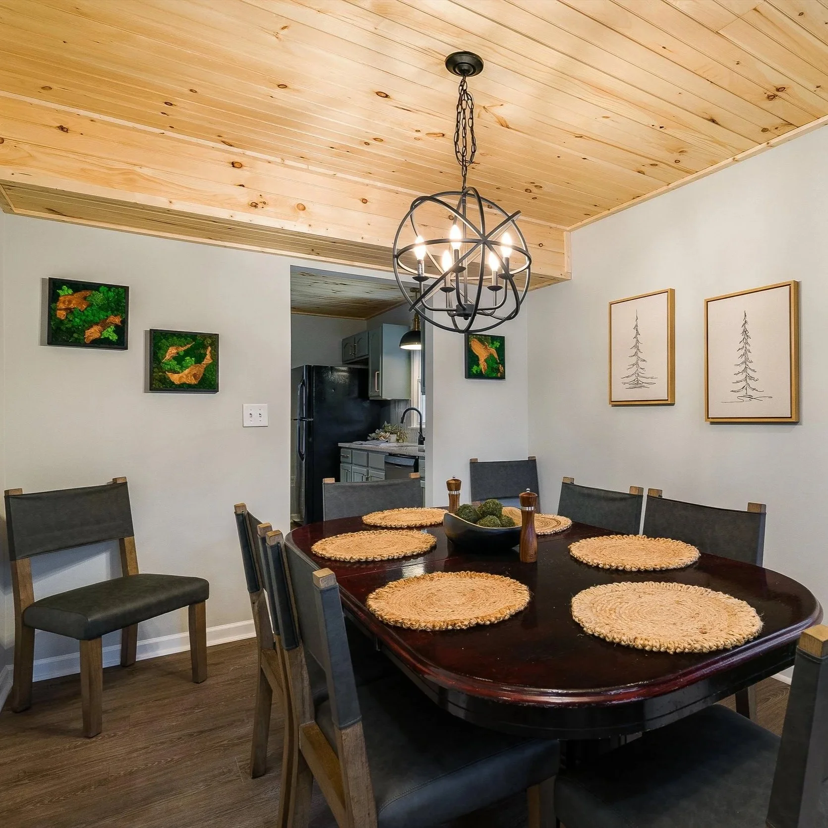 Dining room with a dark wood oval table, six chairs, woven placemats, a black bowl with decorative green balls, and a black chandelier hanging from a wooden ceiling. The walls feature framed artwork of trees and animals. An open doorway leads to a kitchen with gray cabinetry and black refrigerator.